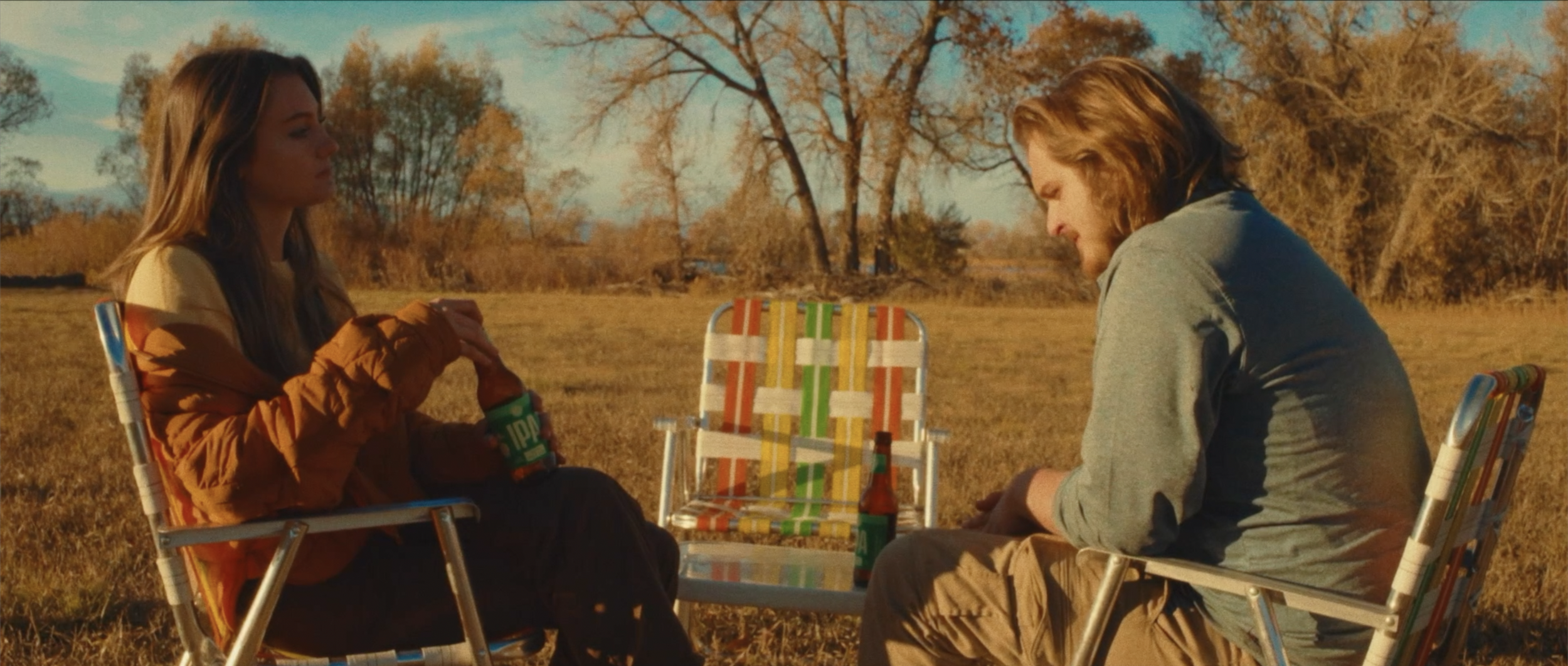 Two young men sitting outdoors in autumn, sitting on colorful lawn chairs, engaging in conversation with beer bottles on a small table between them, in a park with trees and a lake in the background.