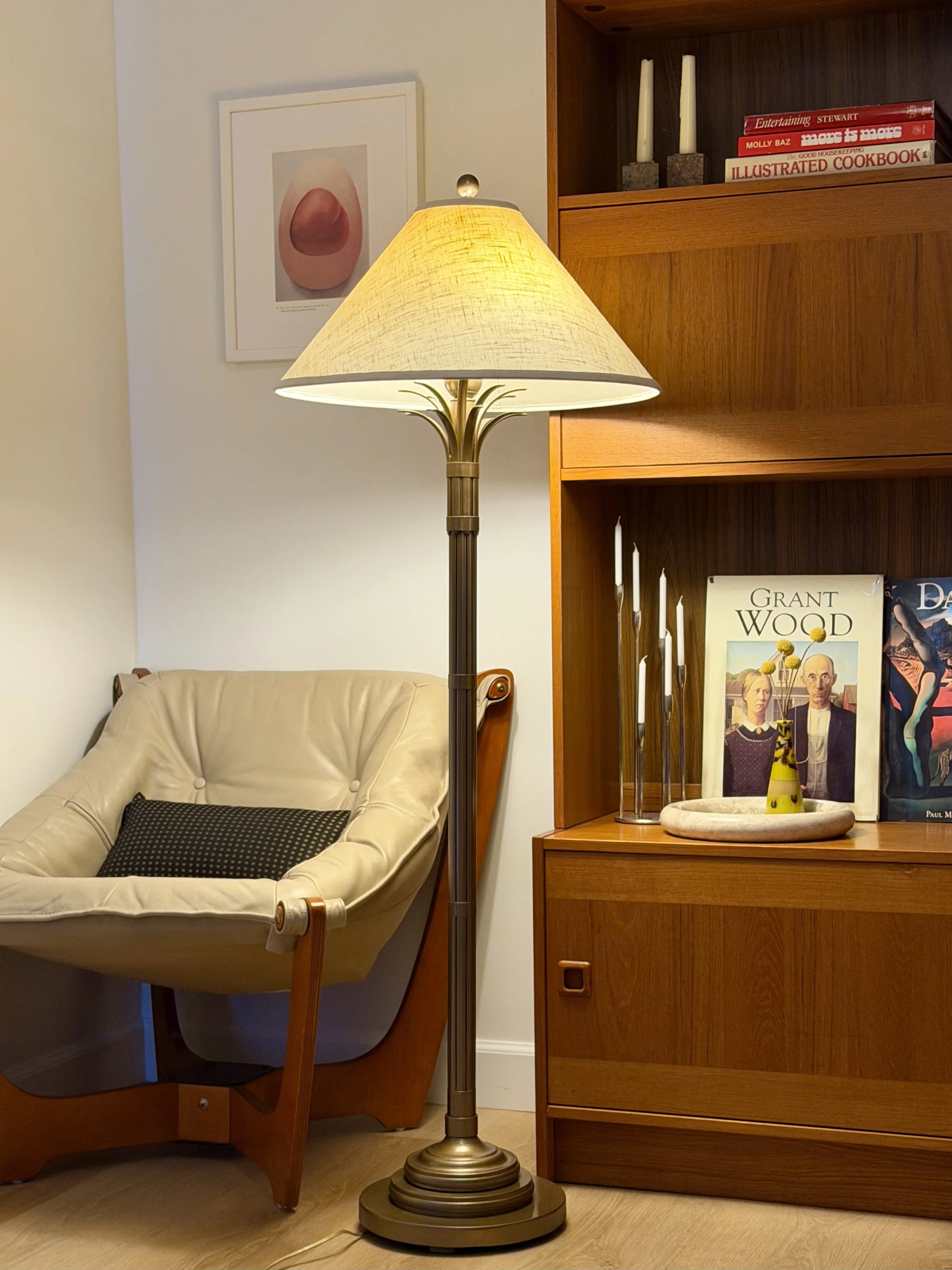 A living room corner with a cream-colored leather armchair, a tall brass floor lamp with a yellow lampshade, and a wooden bookshelf displaying books, candles, and decorative items.