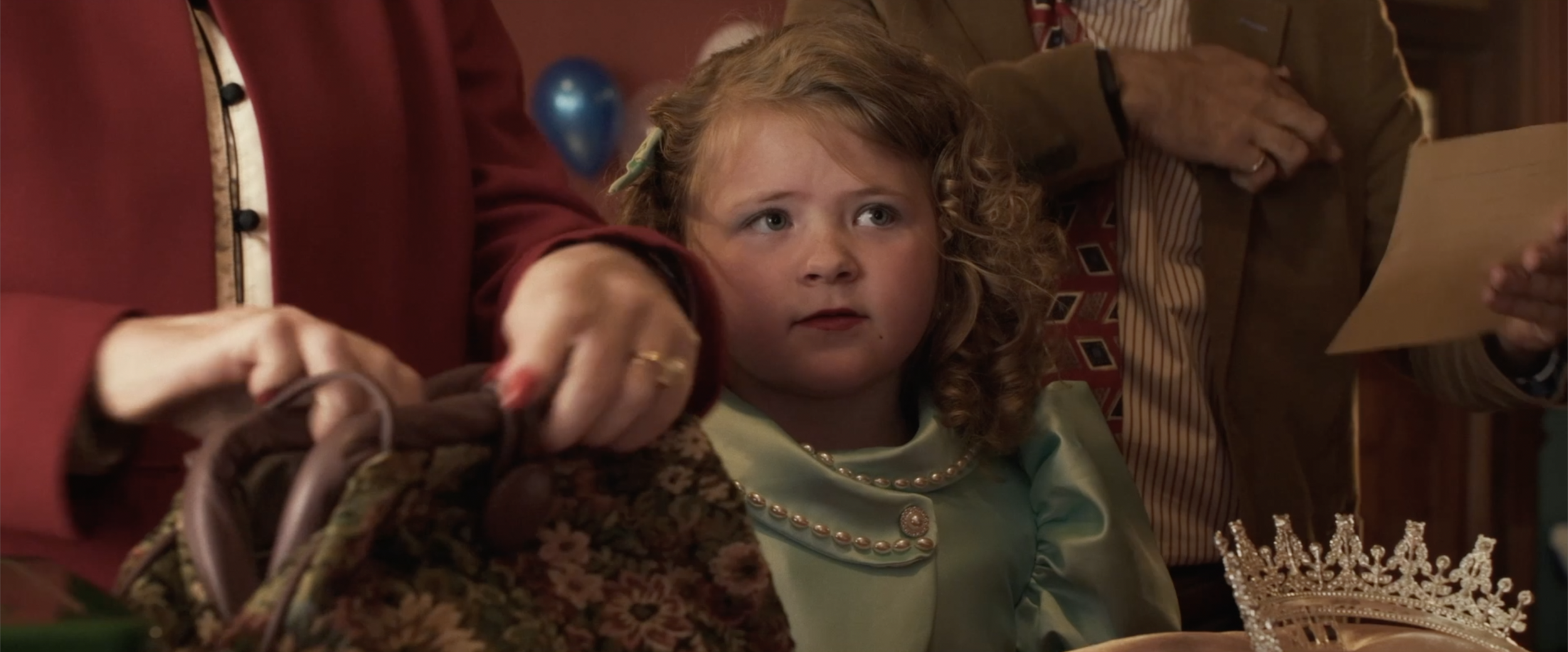 Young girl with curly hair and a pearl necklace sitting at a celebration or party, with a crown on the table in front of her, surrounded by adults.