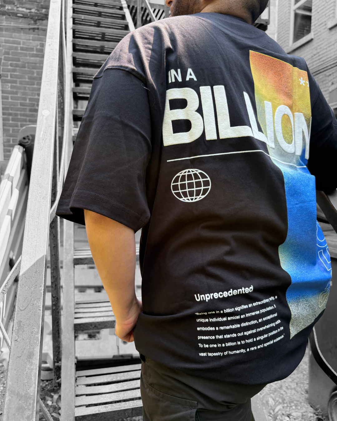 Person standing near a staircase, wearing a black T-shirt with a colorful graphic design and the words 'IN A BILLION' on the back, with some text about being one in a billion.