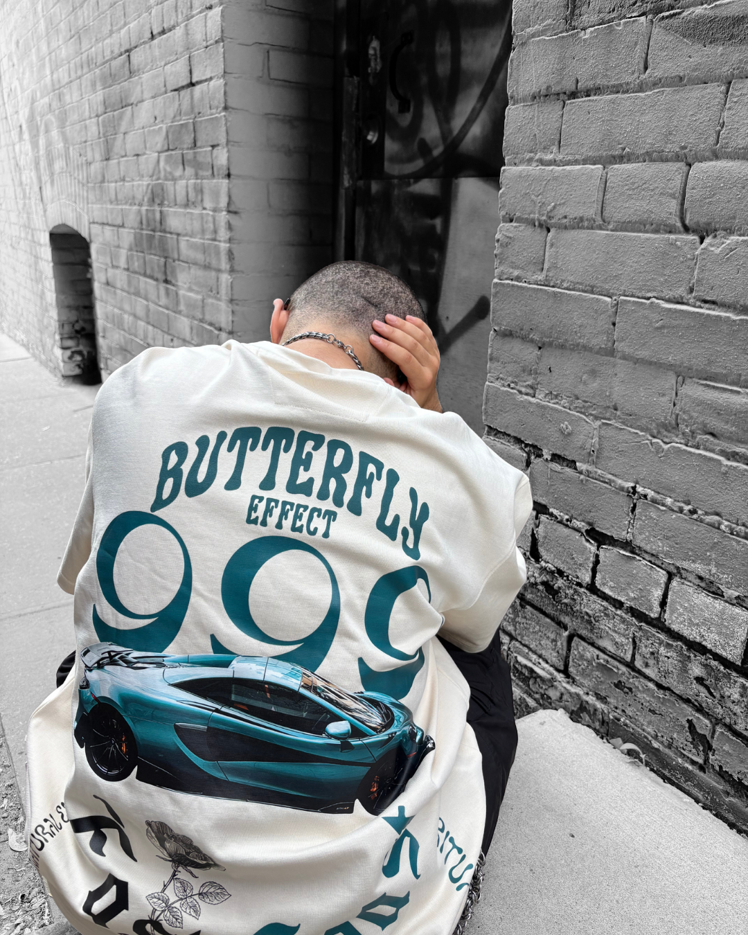 A person with a shaved head and chain necklace is sitting on the ground against a brick wall, holding their head with one hand. They are wearing a white T-shirt with a graphic of a blue sports car and text that reads 'BUTTERFLY EFFECT 99C'.