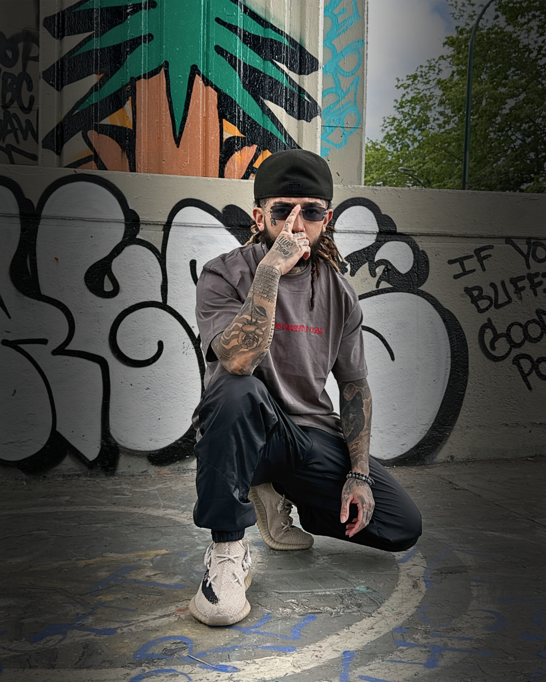 A tattooed man wearing a black cap, gray shirt, black pants, and beige sneakers crouches on graffiti-covered ground and wall, with urban street art and a tree in the background.