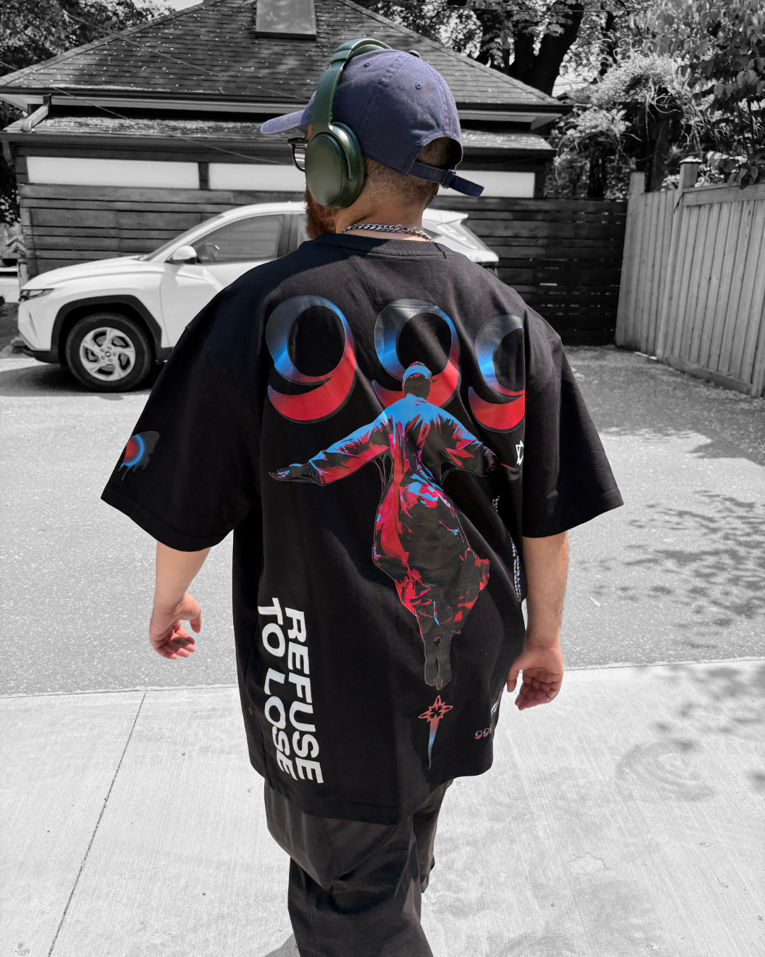 A person with a beard, wearing a blue cap backwards, green headphones, and a black T-shirt with colorful graphics of Jesus on the cross and the words "REFUSE TO LOSE," walking on a sidewalk in a residential area.