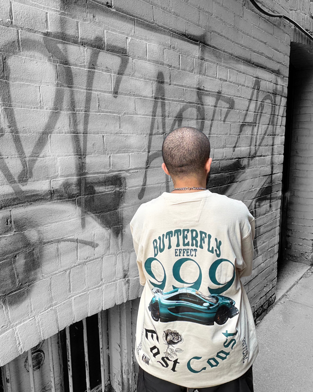 A person with a shaved head and a chain necklace standing with their back to the camera, facing a brick wall with spray-painted graffiti.