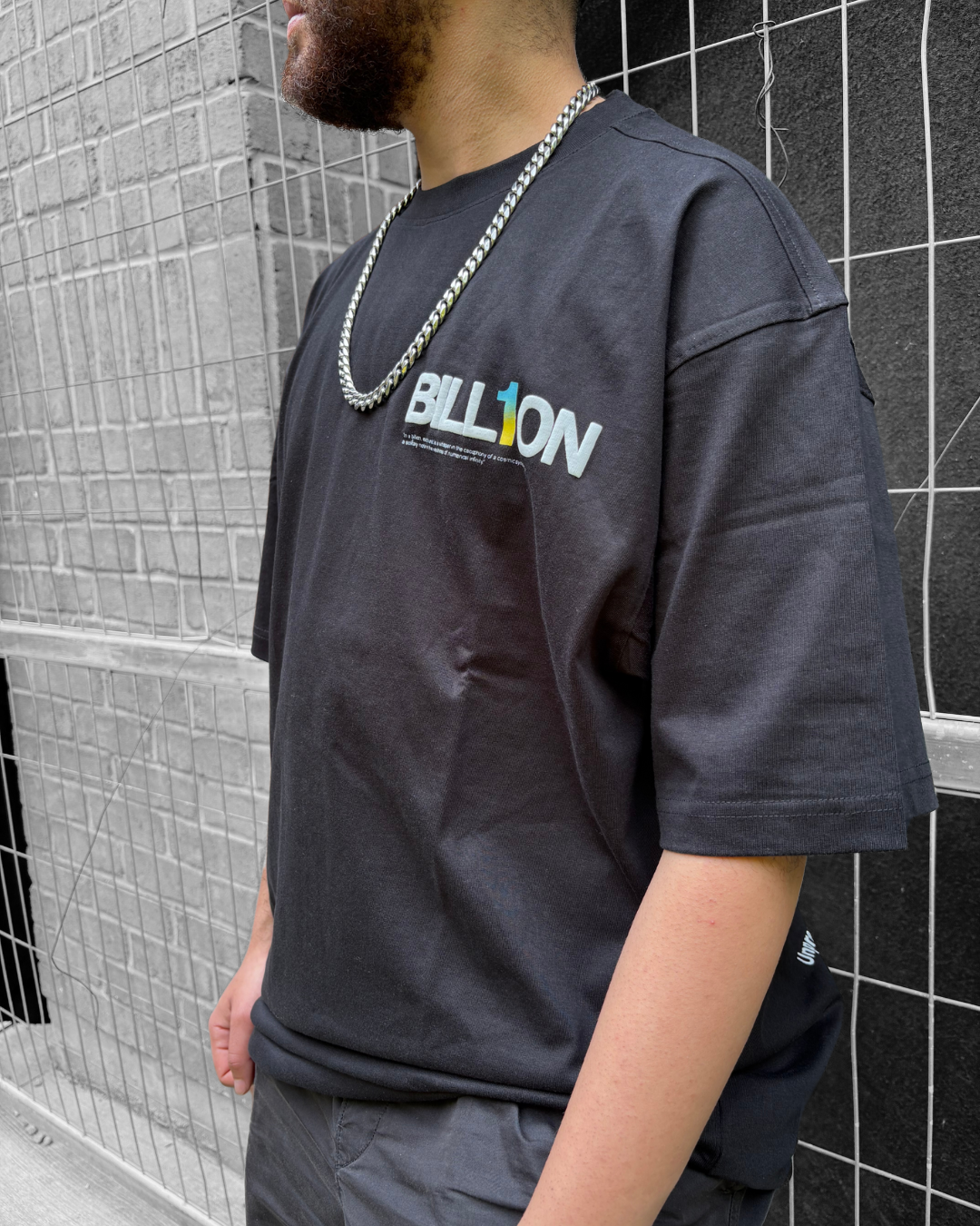Close-up of a person wearing a black T-shirt with colorful embroidered text 'BILLION' and a silver chain necklace, standing against a wire fence and brick wall.