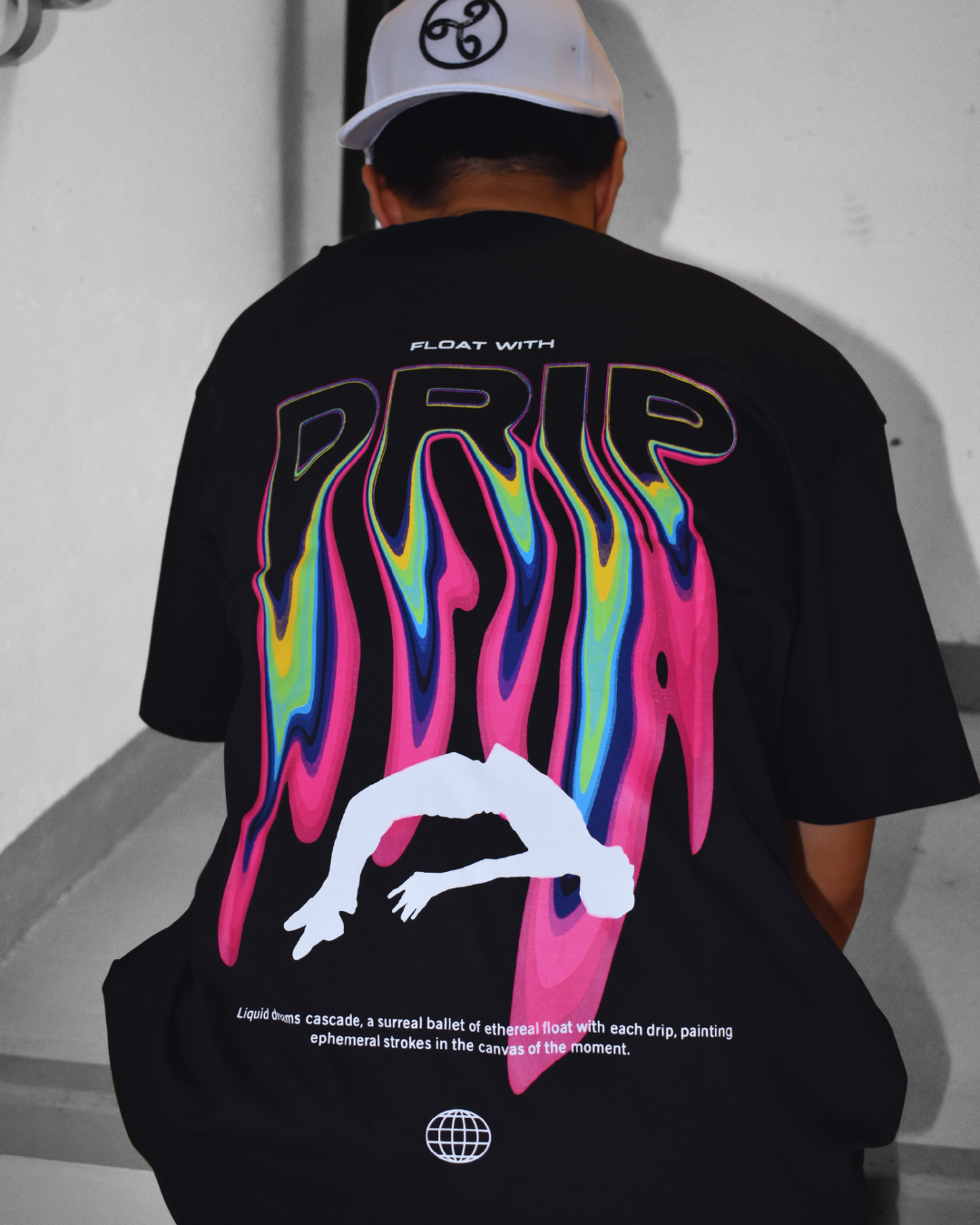 Person wearing a black graphic T-shirt with colorful, wavy patterns and the word 'PRISM' on the back, along with a white cap featuring a logo, standing against a plain wall.