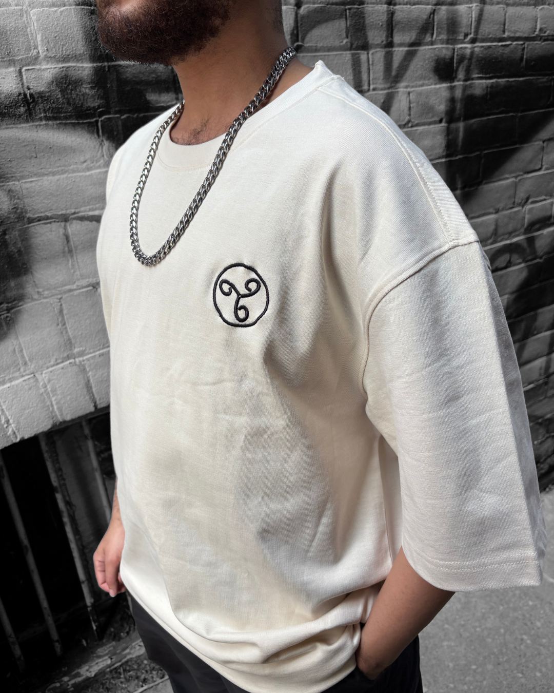 A man wearing a white t-shirt with a black emblem on the left chest, a silver chain necklace, standing against a black and white brick wall.