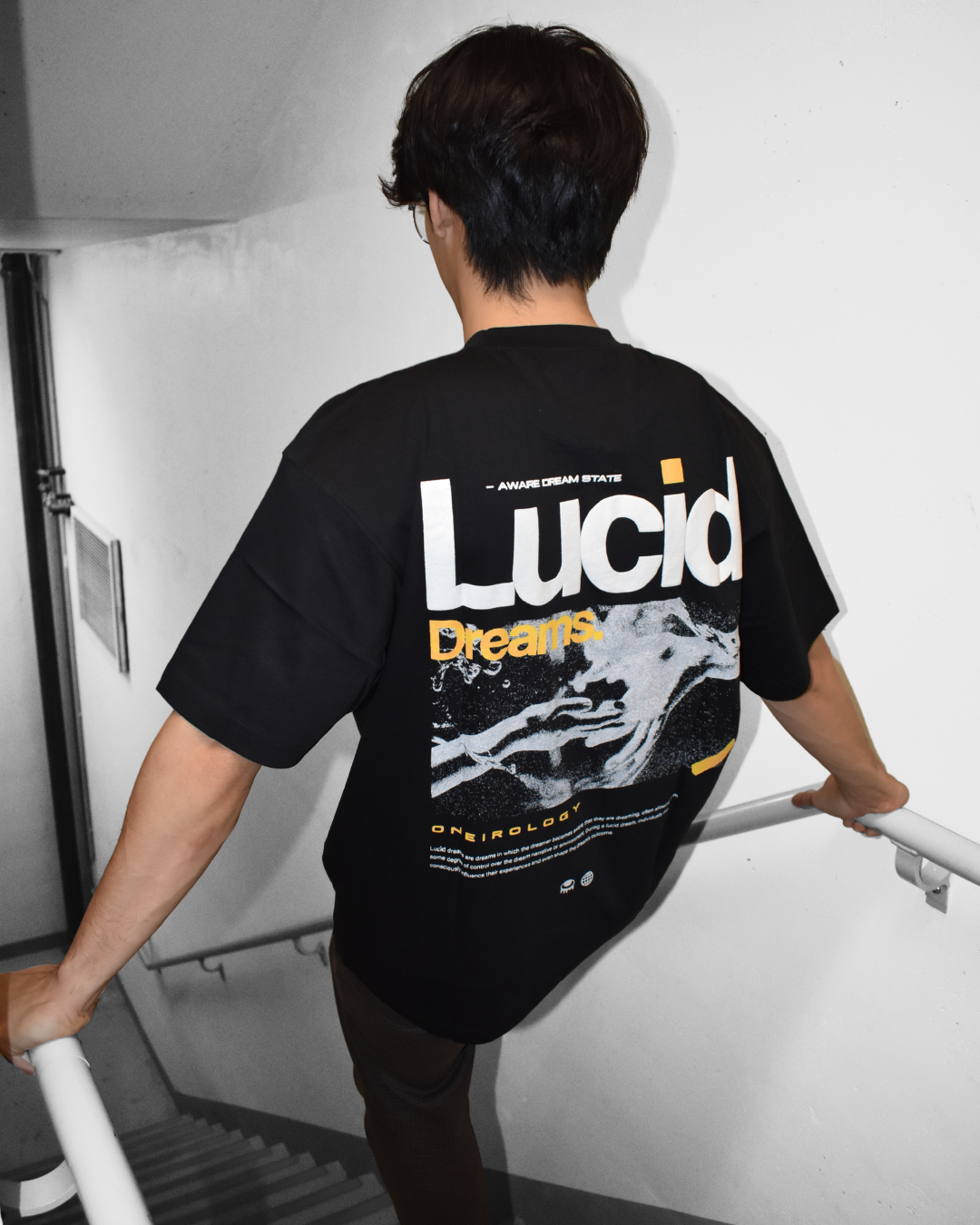 Person walking down a staircase from behind, wearing a black T-shirt with white, yellow, and orange graphic and text including the words "Lucid Dreams" and "Oneirology."