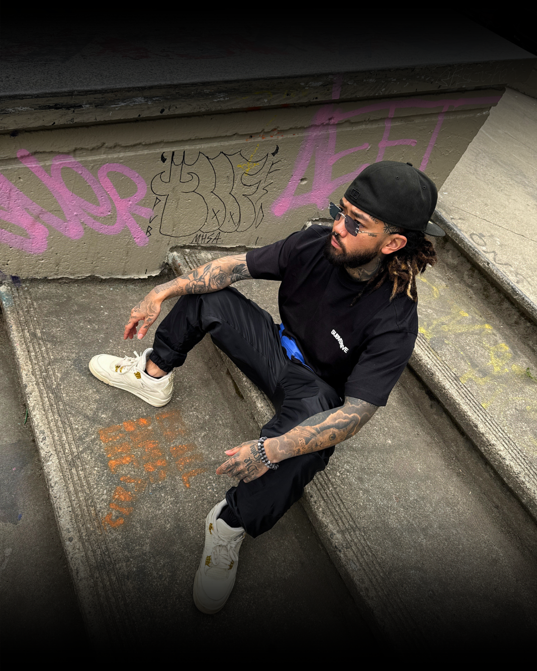 A man with tattoos, wearing sunglasses, a black cap, black shirt, and black pants, sitting on concrete stairs in front of a graffiti-covered wall.