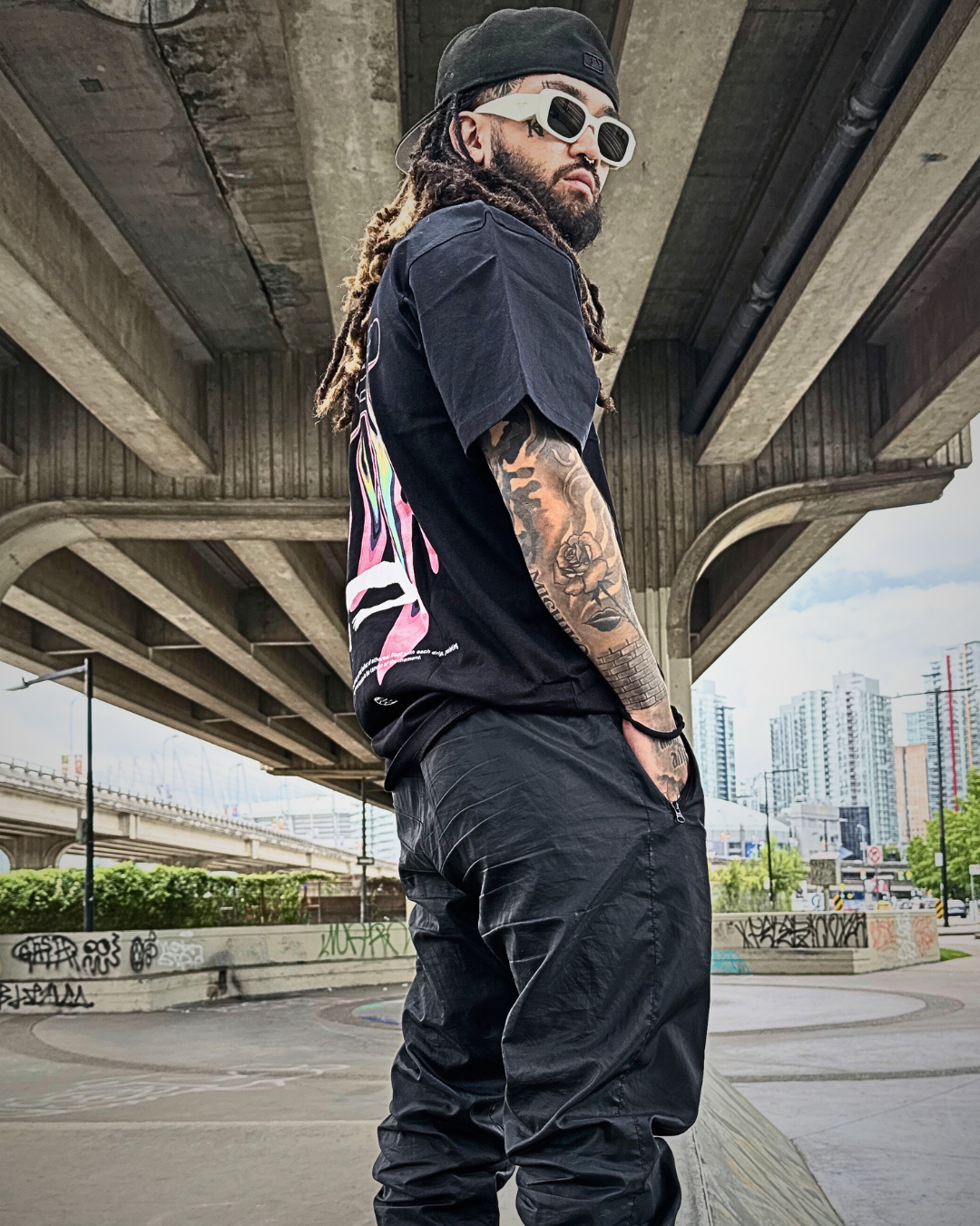 A man with long dreadlocks, wearing sunglasses, a black cap backwards, a black graphic t-shirt, and black pants, standing under a large concrete bridge with an urban cityscape in the background.