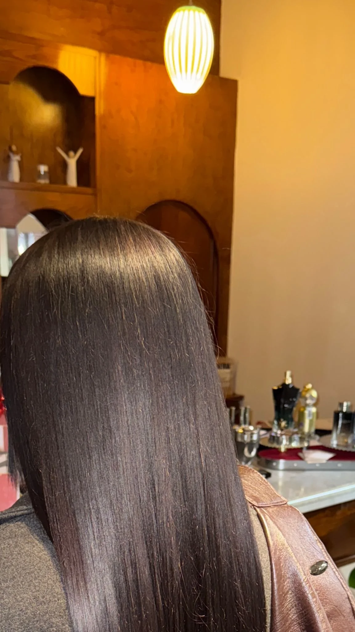 Close-up of a woman with straight, shiny, brown hair indoors, with a wooden wall, decorative figures, and a warm-colored lamp in the background.