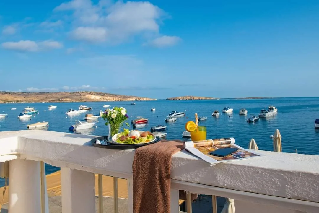 A seaside patio with a view of boats floating on calm water, a vase with flowers, a plate of food, a glass of yellow drink, newspapers, and magazines on a white balcony railing.