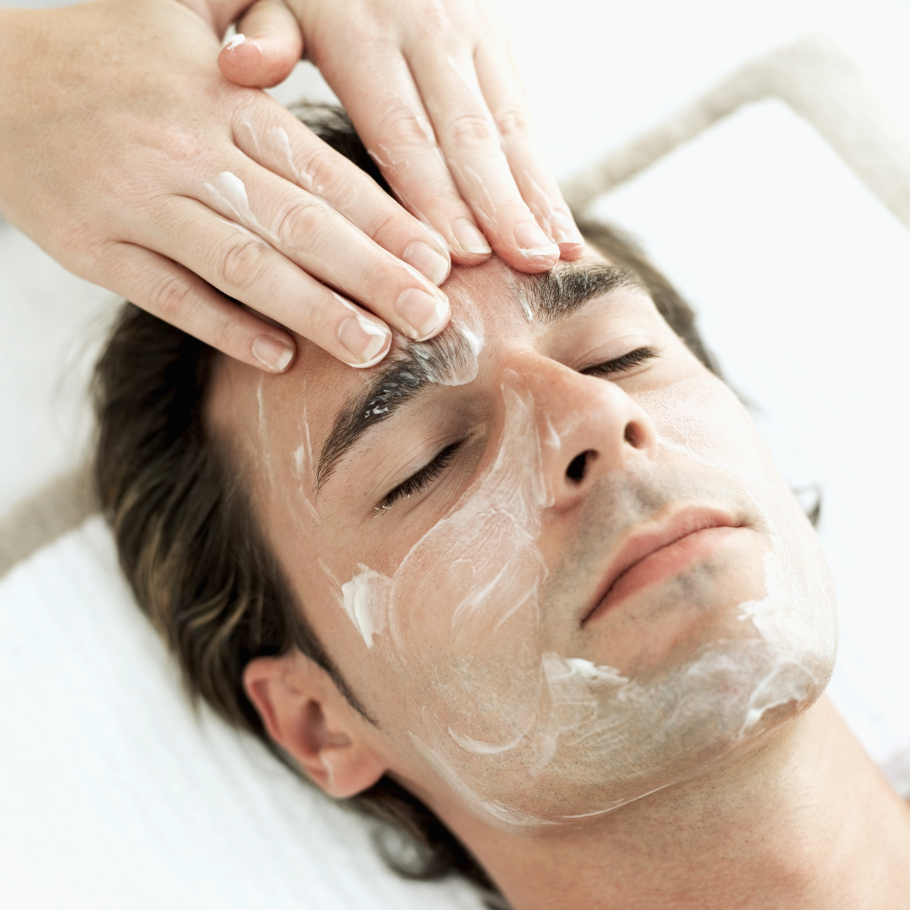 A man receives a facial treatment, with a therapist applying a cream or mask on his face.