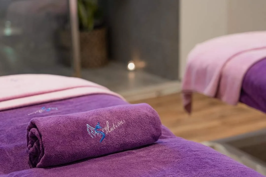 Two massage tables with purple towels, one with an embroidered logo of a hummingbird and the word 'Baluluna', in a spa or wellness room.