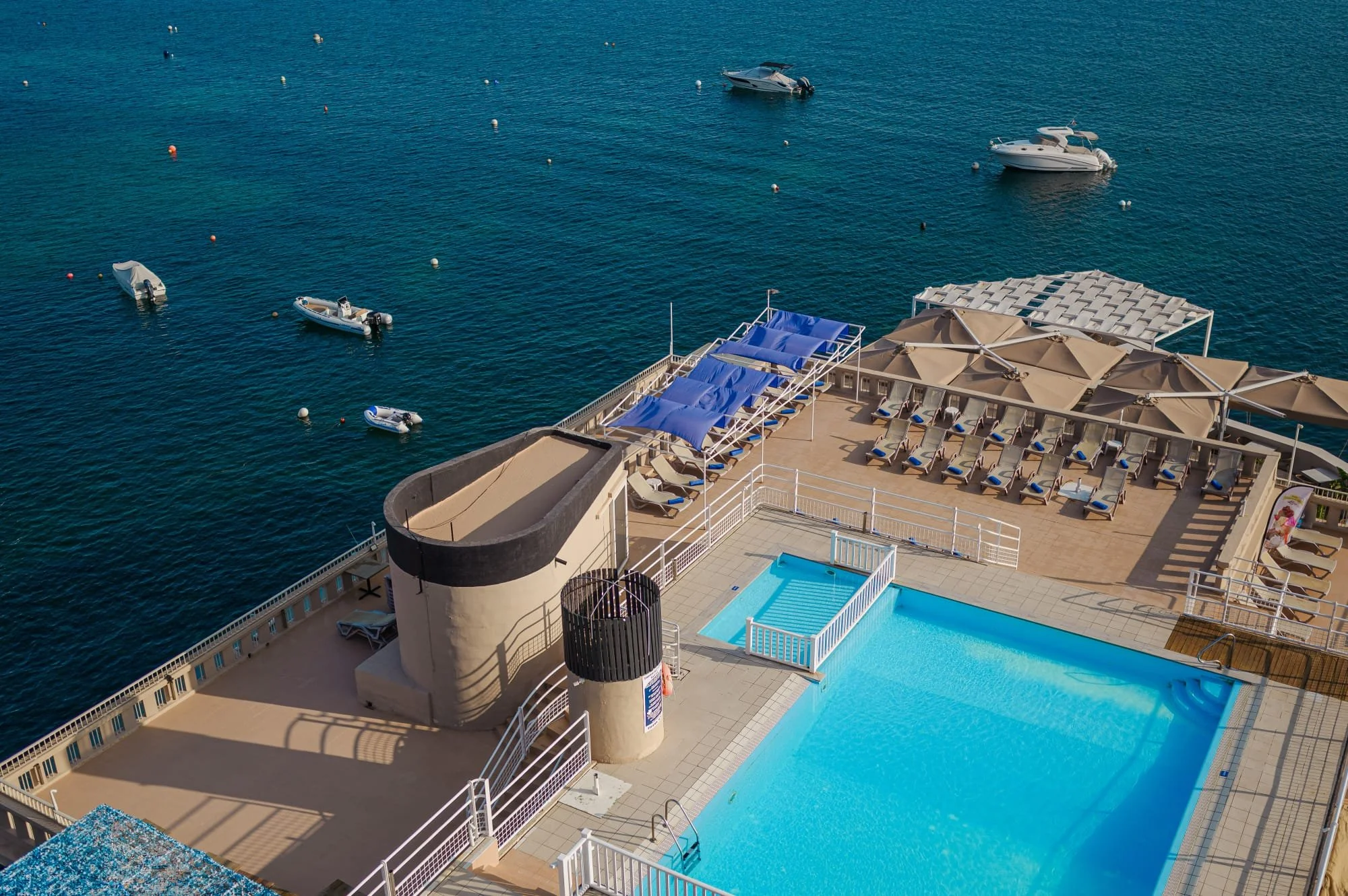Aerial view of a hotel rooftop with swimming pool, lounge chairs, umbrellas, and boats in the water.