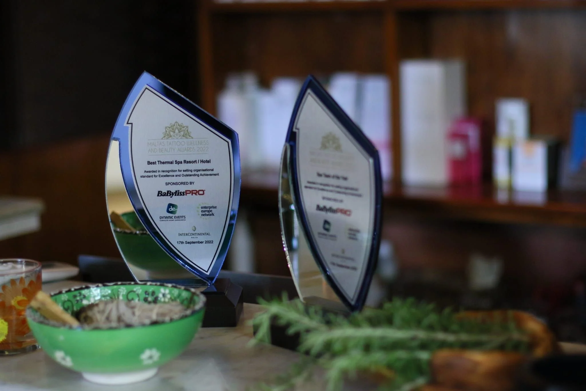 Two glass awards on a table, one recognizing a spa as the best thermal spa resort or hotel, with the date 17th September 2022, and various food and drink items and a plant in the foreground.