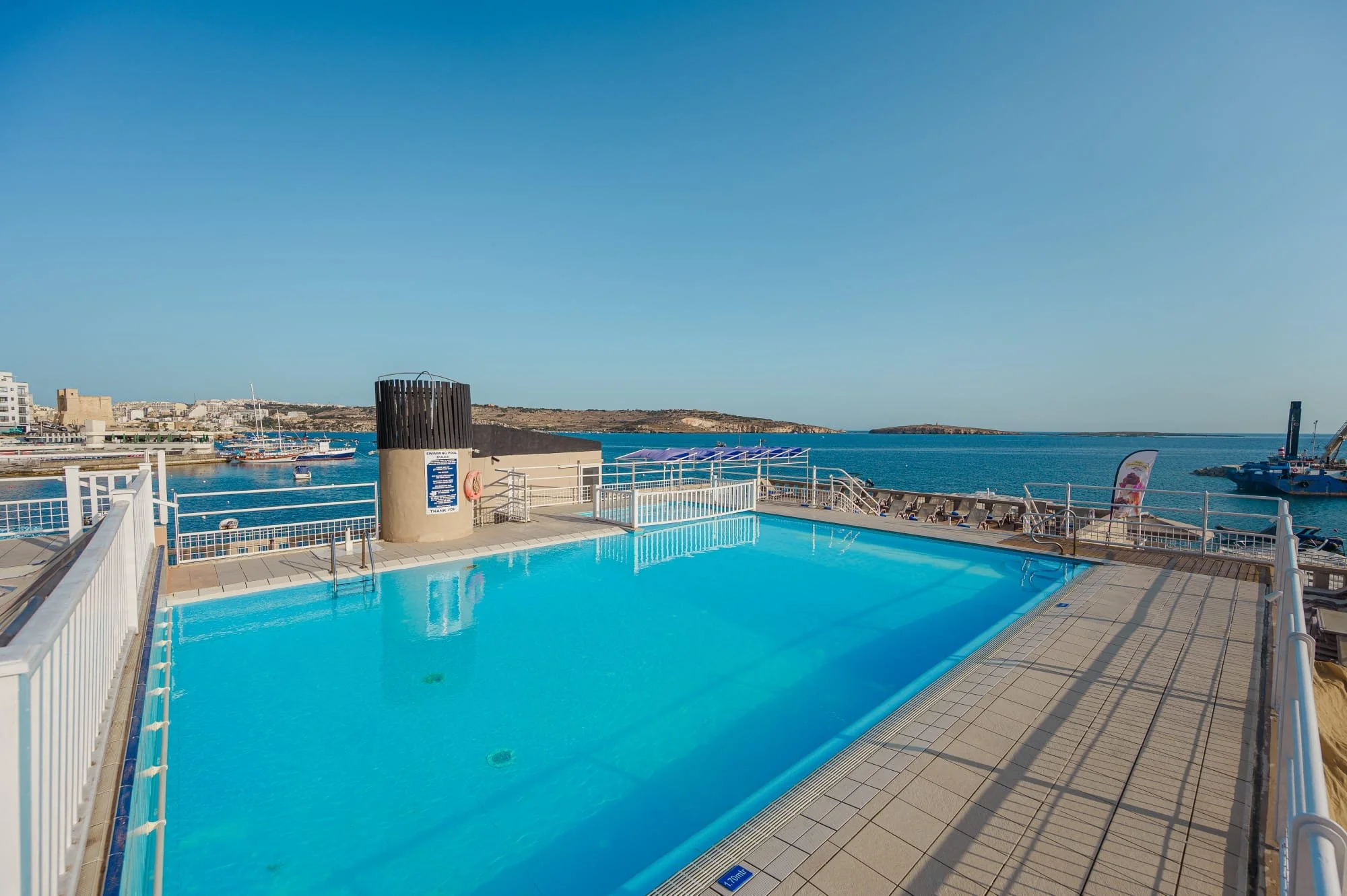 Swimming pool overlooking the ocean with boats, deck chairs, and a distant island on a clear day.