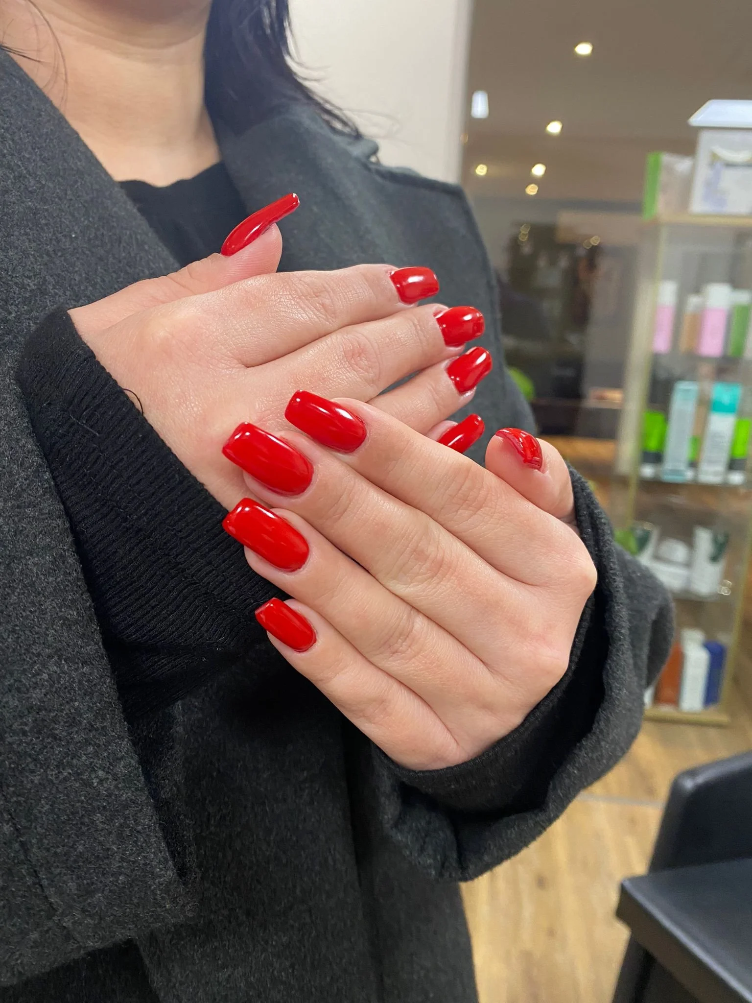 Close-up of a woman with red painted nails, wearing a dark coat, with her hands crossed over her chest, in an indoor setting that appears to be a store or pharmacy.