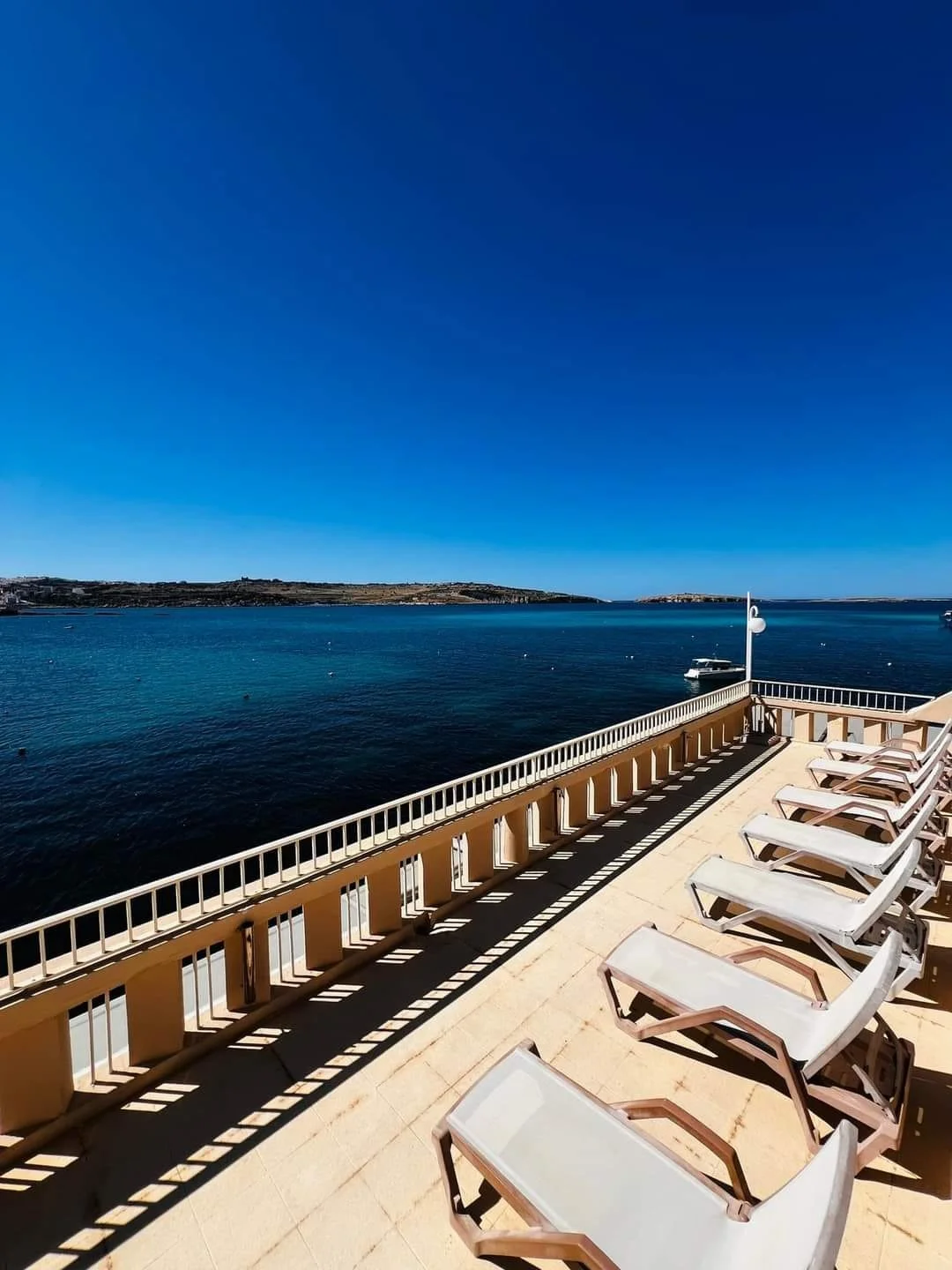 Poolside lounge chairs on a deck overlooking a calm ocean with a boat and distant land under a clear blue sky.