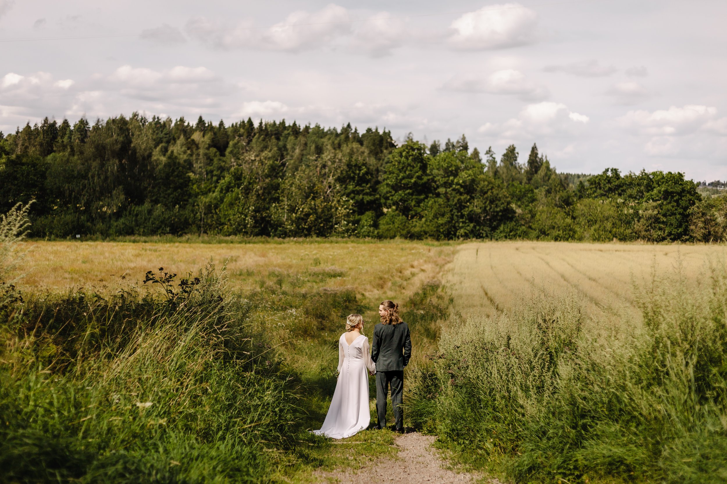 Häätapahtuma maaseudulla, morsian ja sulhanen kävelevät polulla, taustalla metsää ja pilviä.
