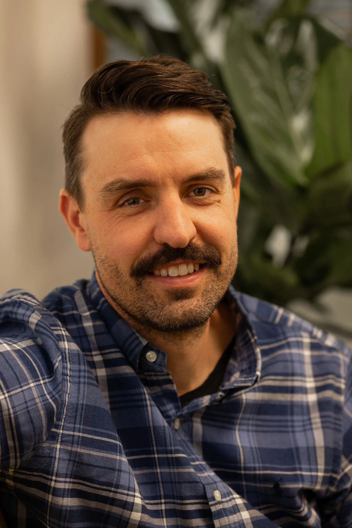 Portrait of a man with brown hair, mustache, wearing a black blazer over a plaid shirt, against a white background.