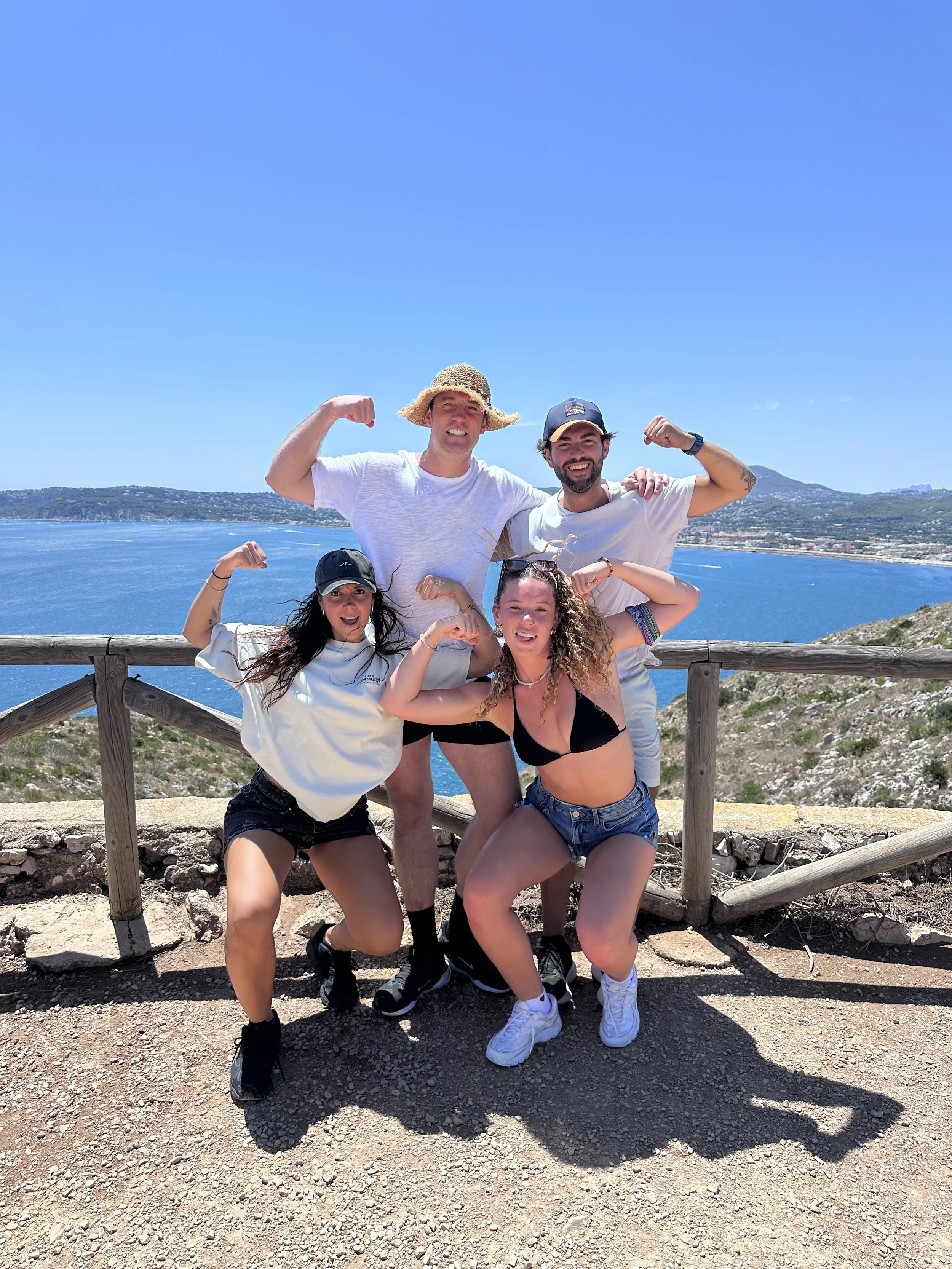 Group of five friends posing on a mountain overlook with a lake and coastline in the background, flexing their arms, wearing casual summer clothes and hats on a sunny day.