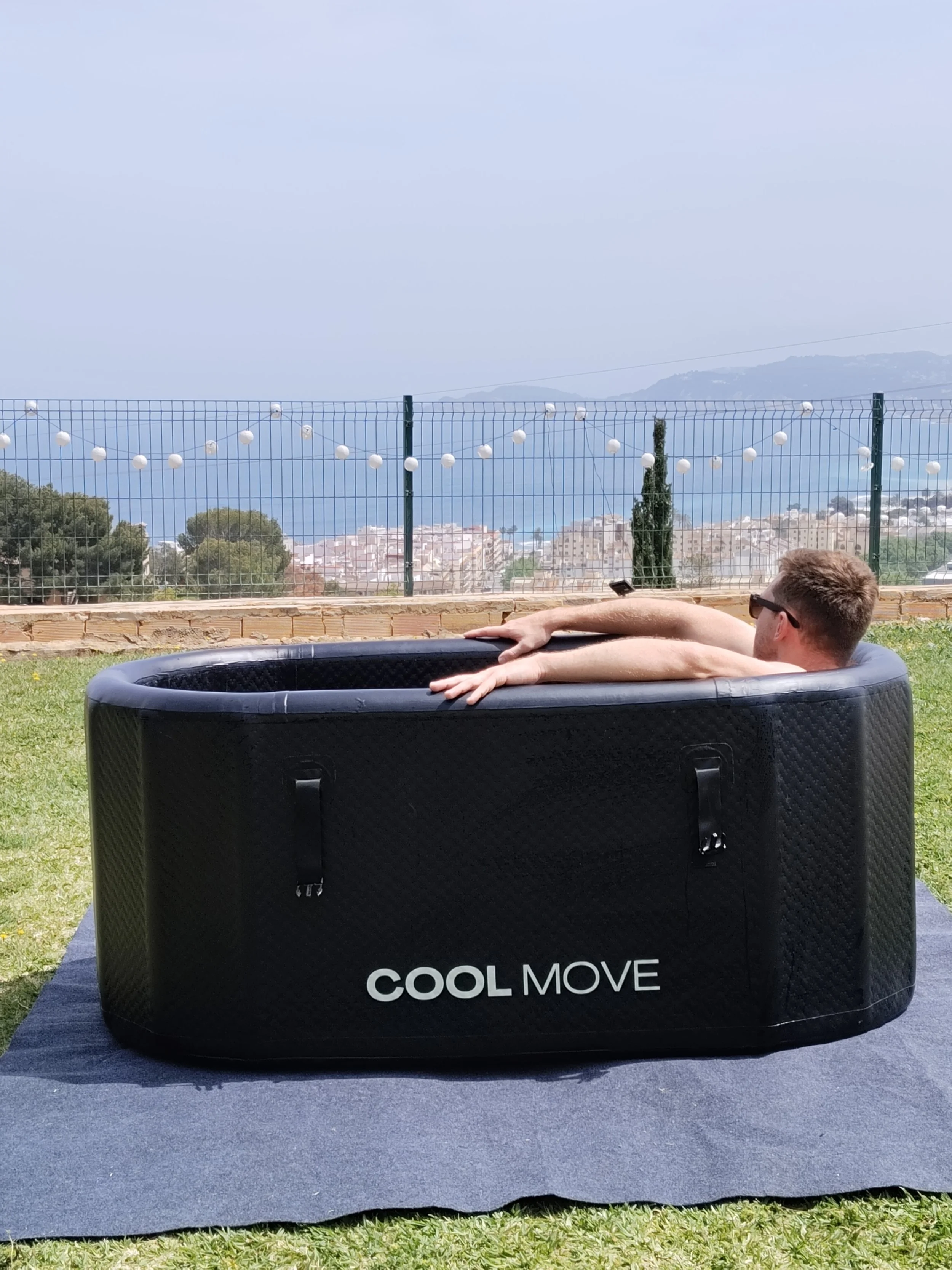 A man in sunglasses relaxing in a black circular portable hot tub labeled 'Cool Move' outdoors on a grassy area with a view of the sea, trees, and a city in the distance, with string lights hanging in the background.