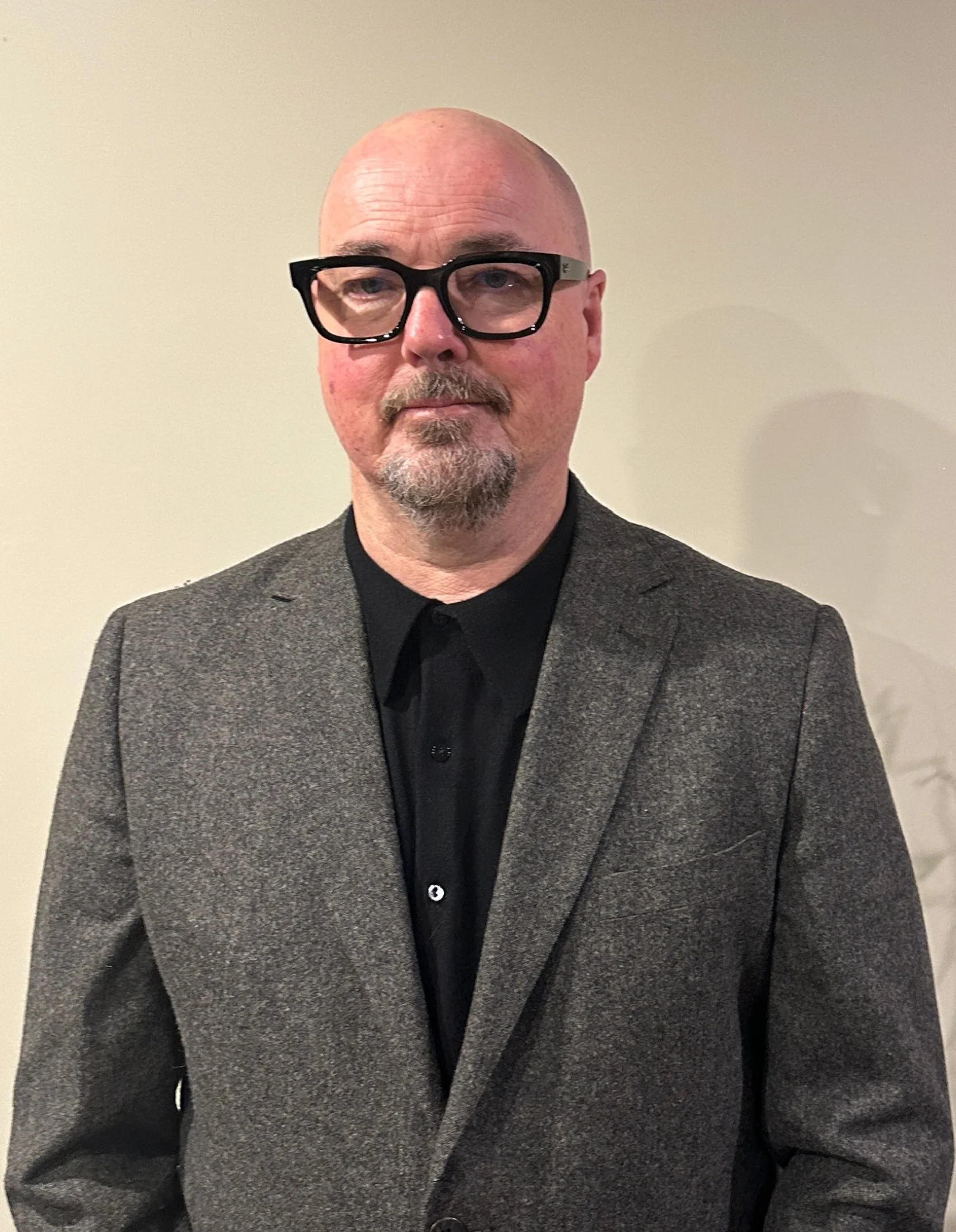 A middle-aged man with a bald head, glasses, and a beard, wearing a dark shirt and a gray blazer, standing against a plain beige wall.