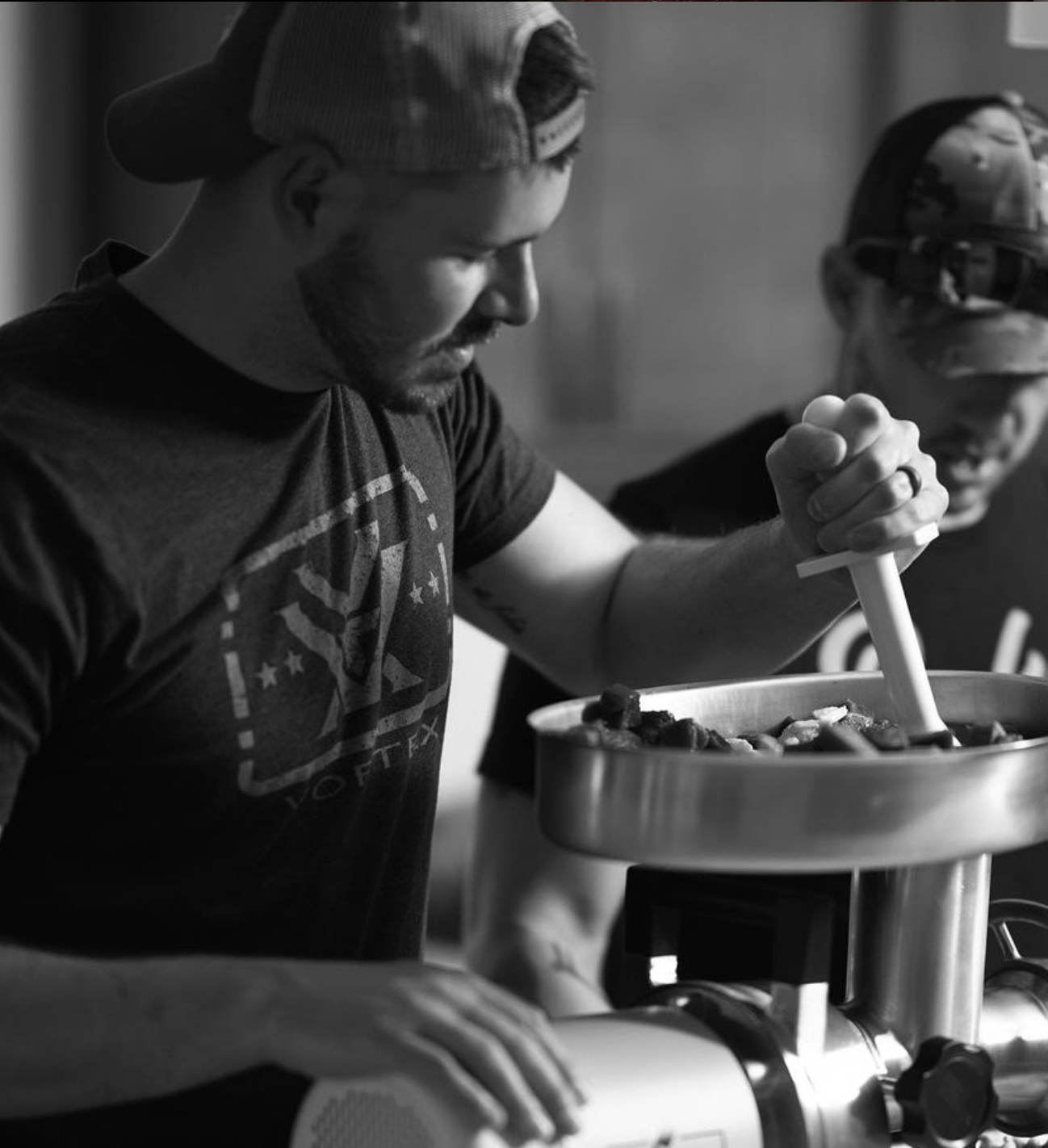 A man wearing a cap and a T-shirt, with a beard, is operating a meat grinder, with another man in military gear and helmet watching in the background.