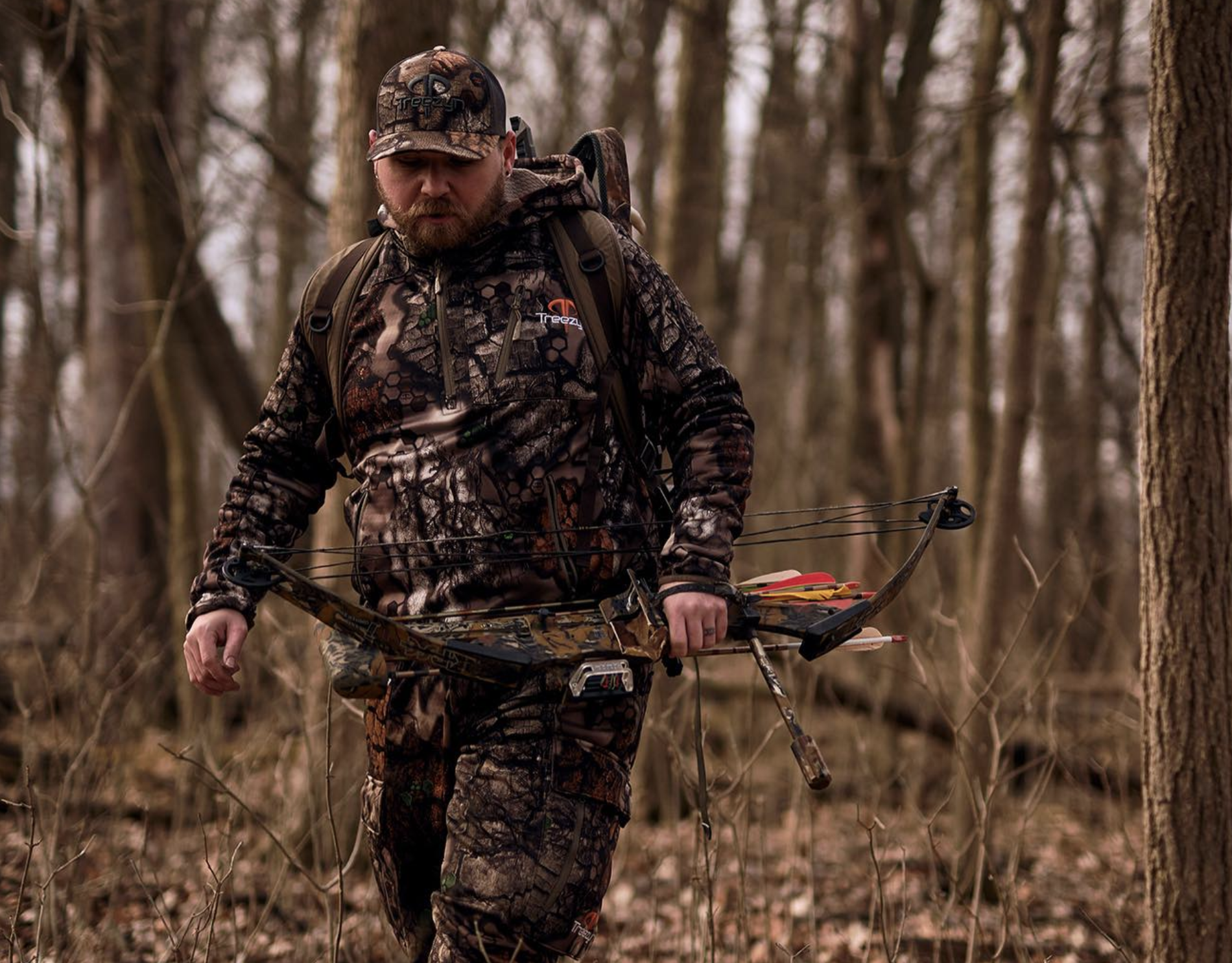 A man in camouflage clothing walking through a leafless forest, carrying a compound bow with arrows attached.