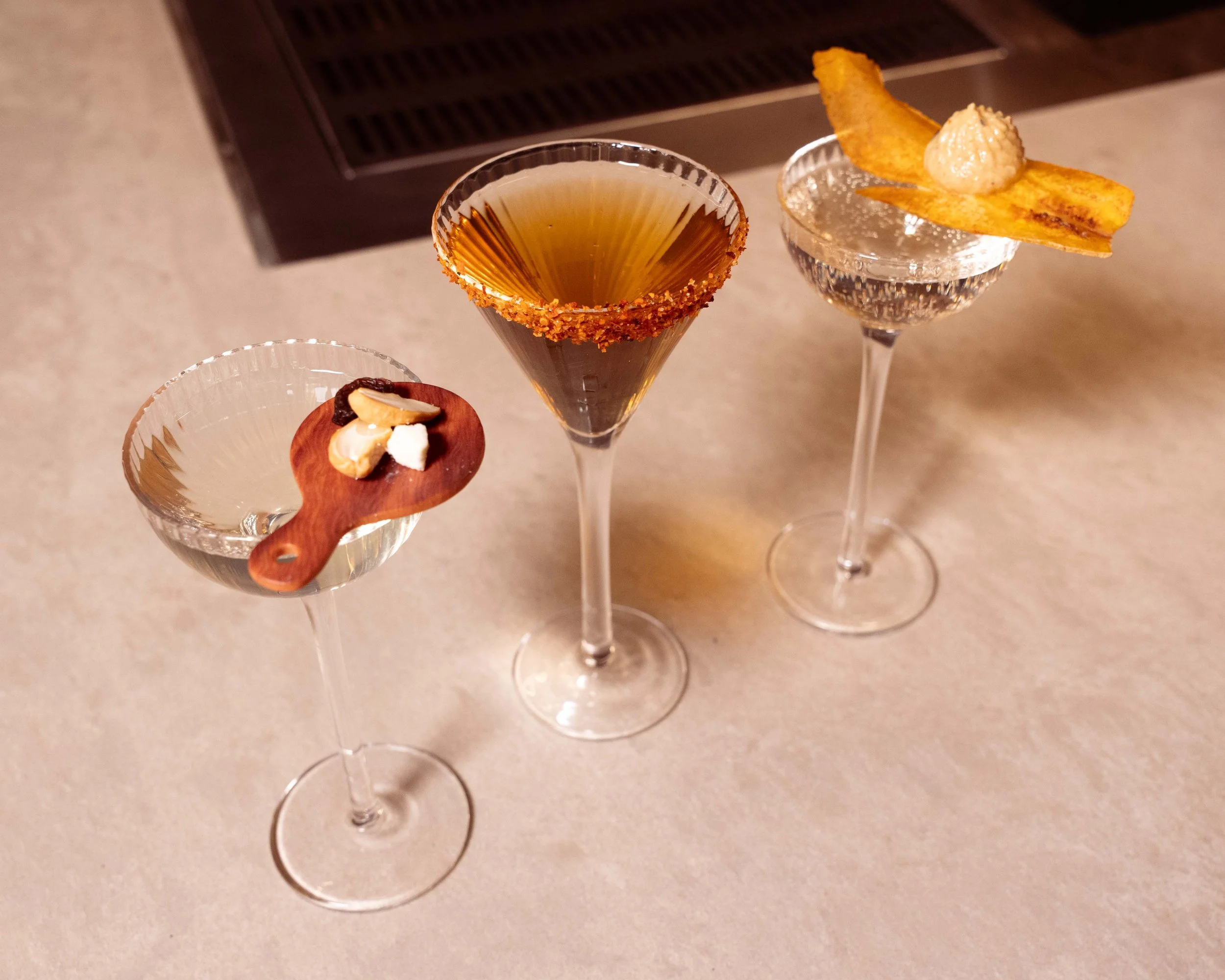 Three cocktail glasses with different drinks and garnishes on a beige countertop. The left glass has a small wooden board with nuts and dried fruit. The middle glass has a dark drink with a chili salt rim. The right glass contains a clear drink with a potato chip and dollop of creamy substance as garnish.