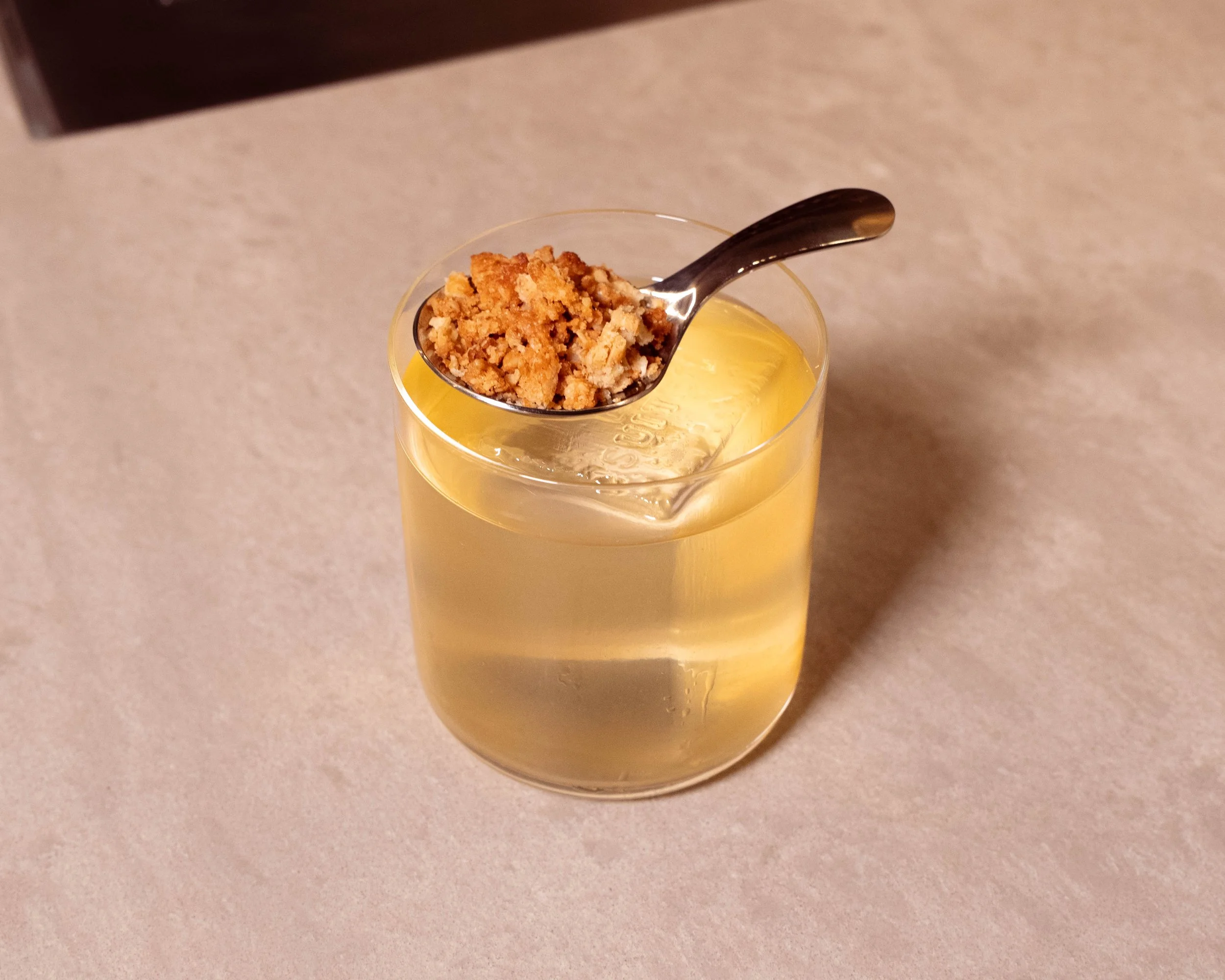 A glass of yellow-colored beverage with a large ice cube, garnished with a silver spoon holding a crumbly brown topping.