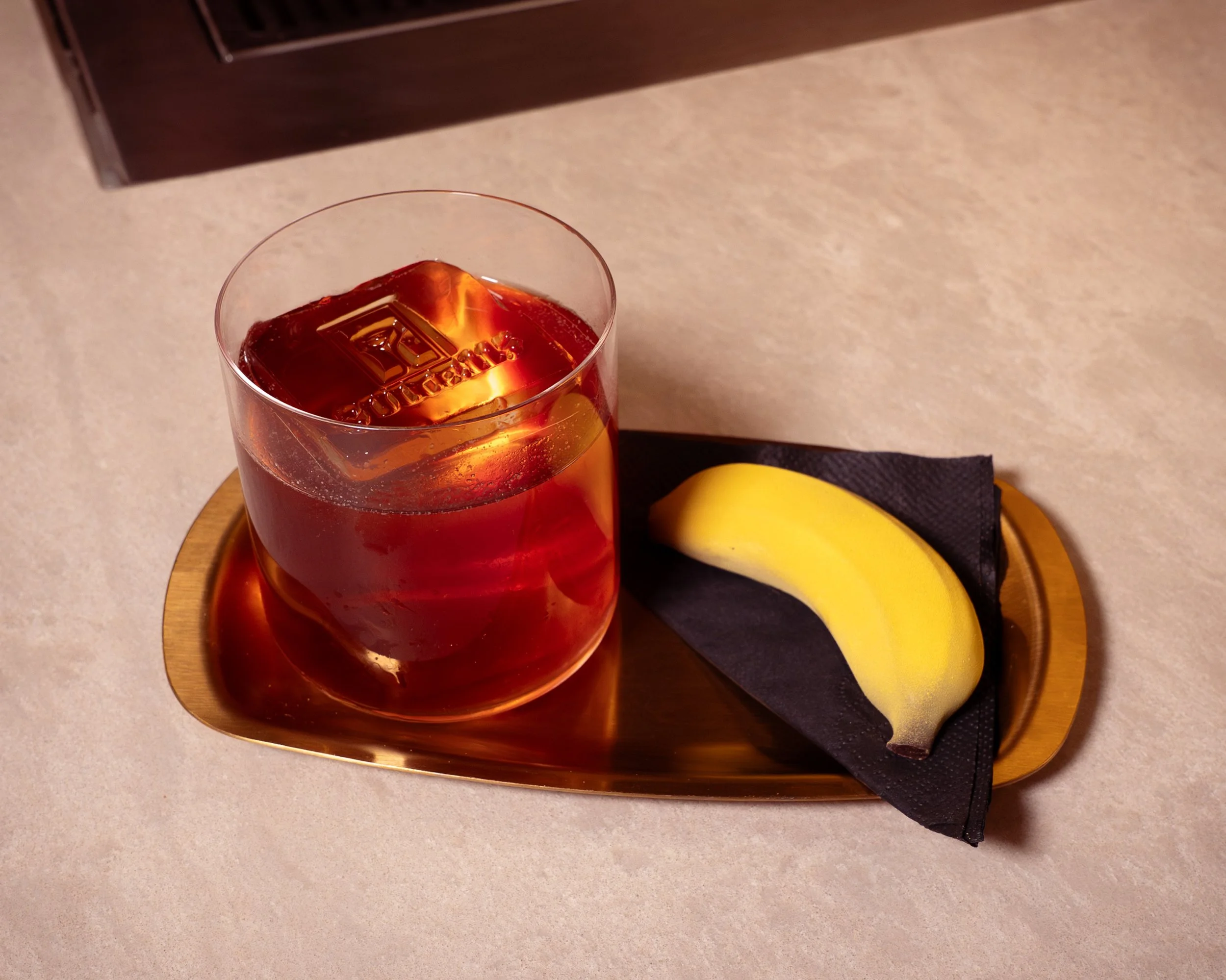 A glass of iced tea with a large ice cube on a gold tray, accompanied by a yellow banana resting on a black napkin.