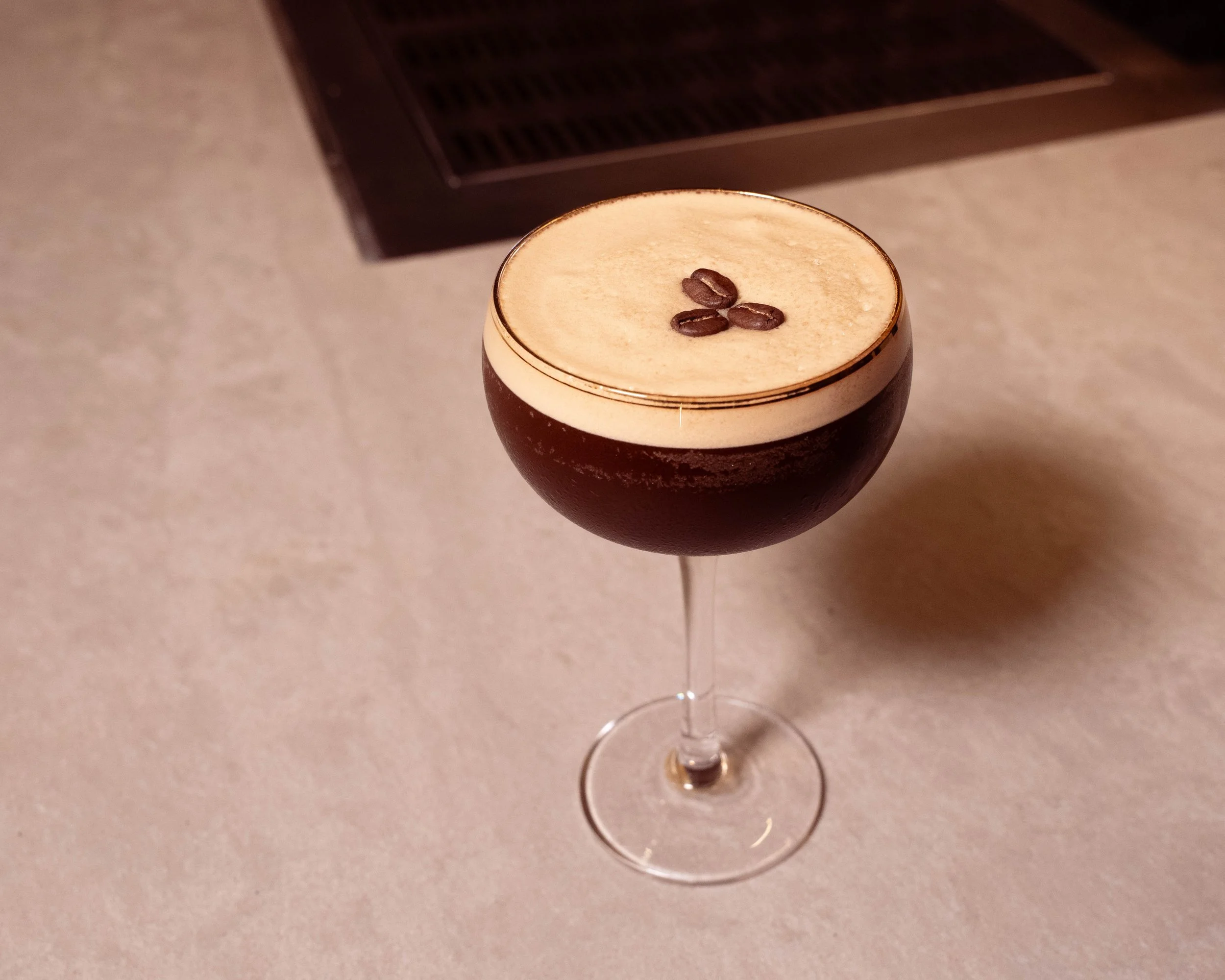 A dark coffee cocktail topped with foam and coffee beans in a coupe glass placed on a light-colored surface.