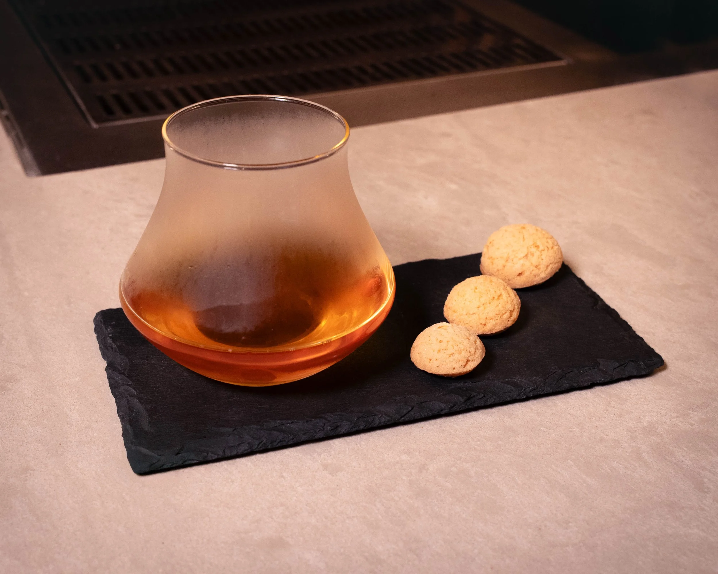 A glass of amber liquor and three small cookies on a black slate coaster on a light-colored countertop.