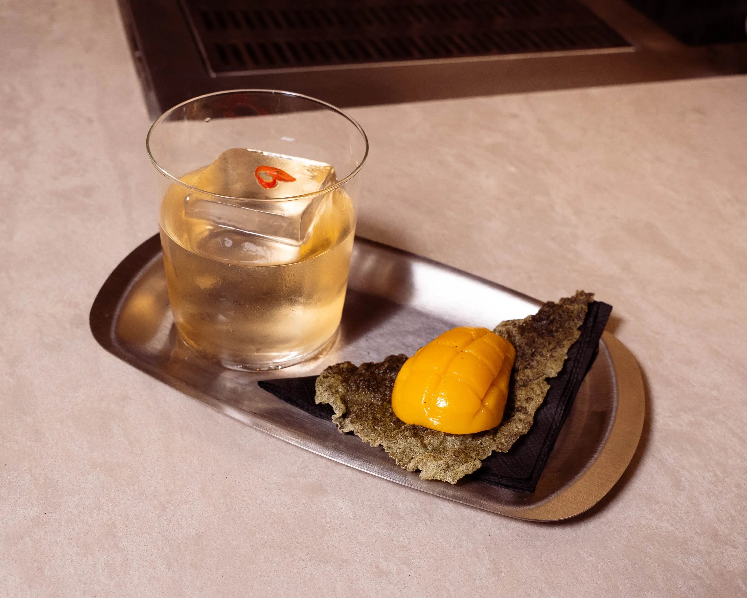 A glass of a clear drink with ice and a red chili pepper on top, next to a rectangular piece of fried food topped with a yellow sauce, all on a metal tray with black napkin on a beige countertop.