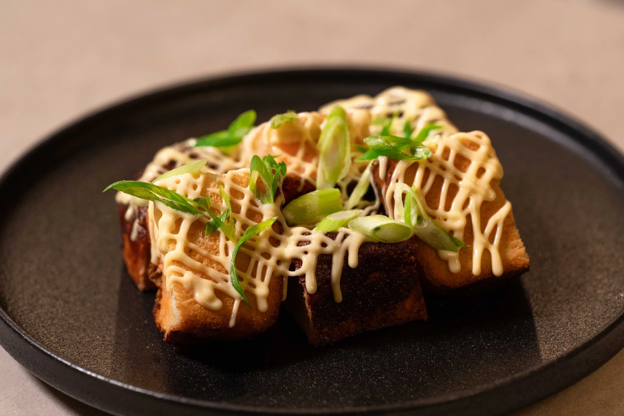 Fried tofu topped with chopped green onions and drizzled with mayonnaise on a black plate.
