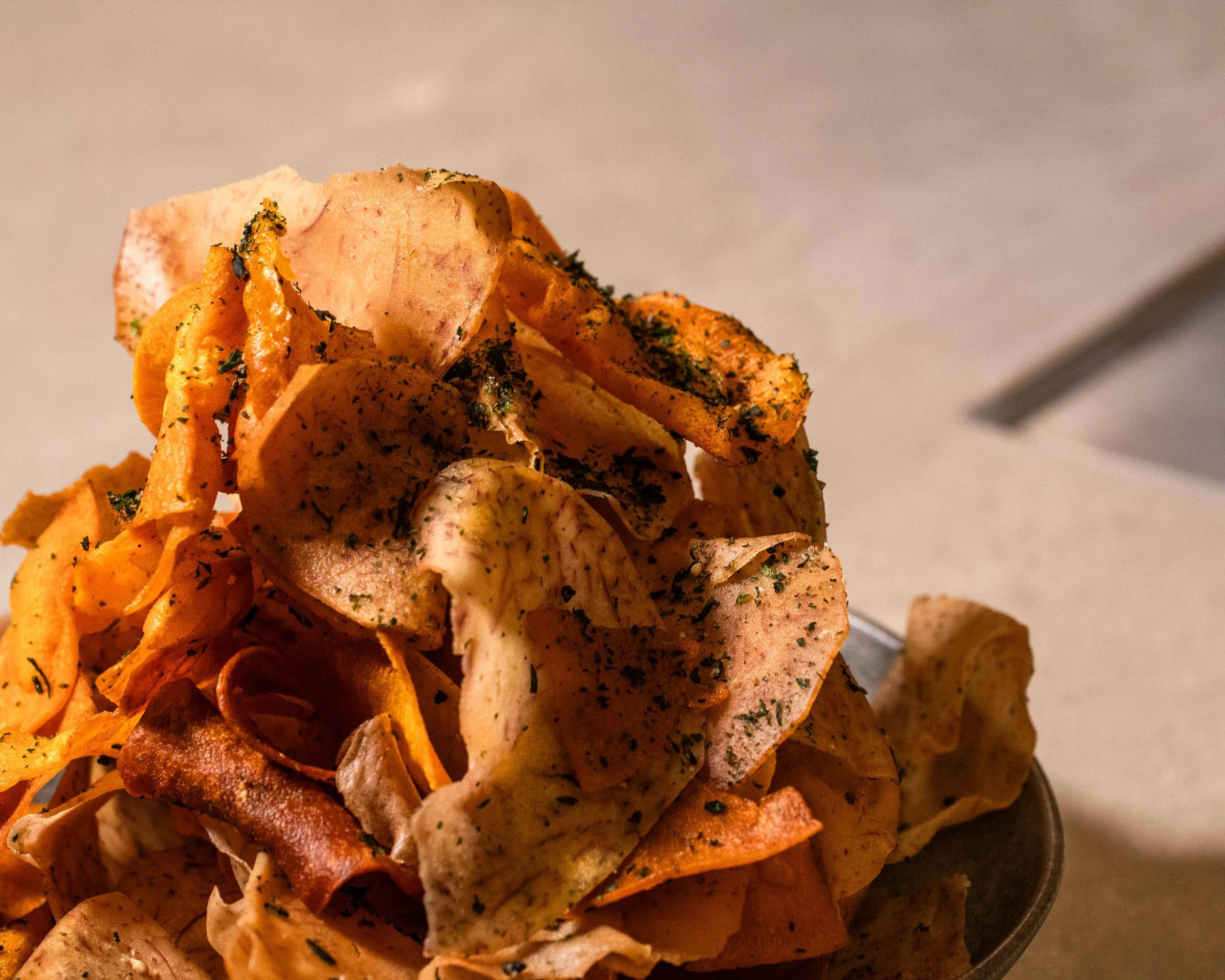 Close-up of a bowl filled with seasoned, crispy potato chips sprinkled with herbs, with a plain, light-colored background.