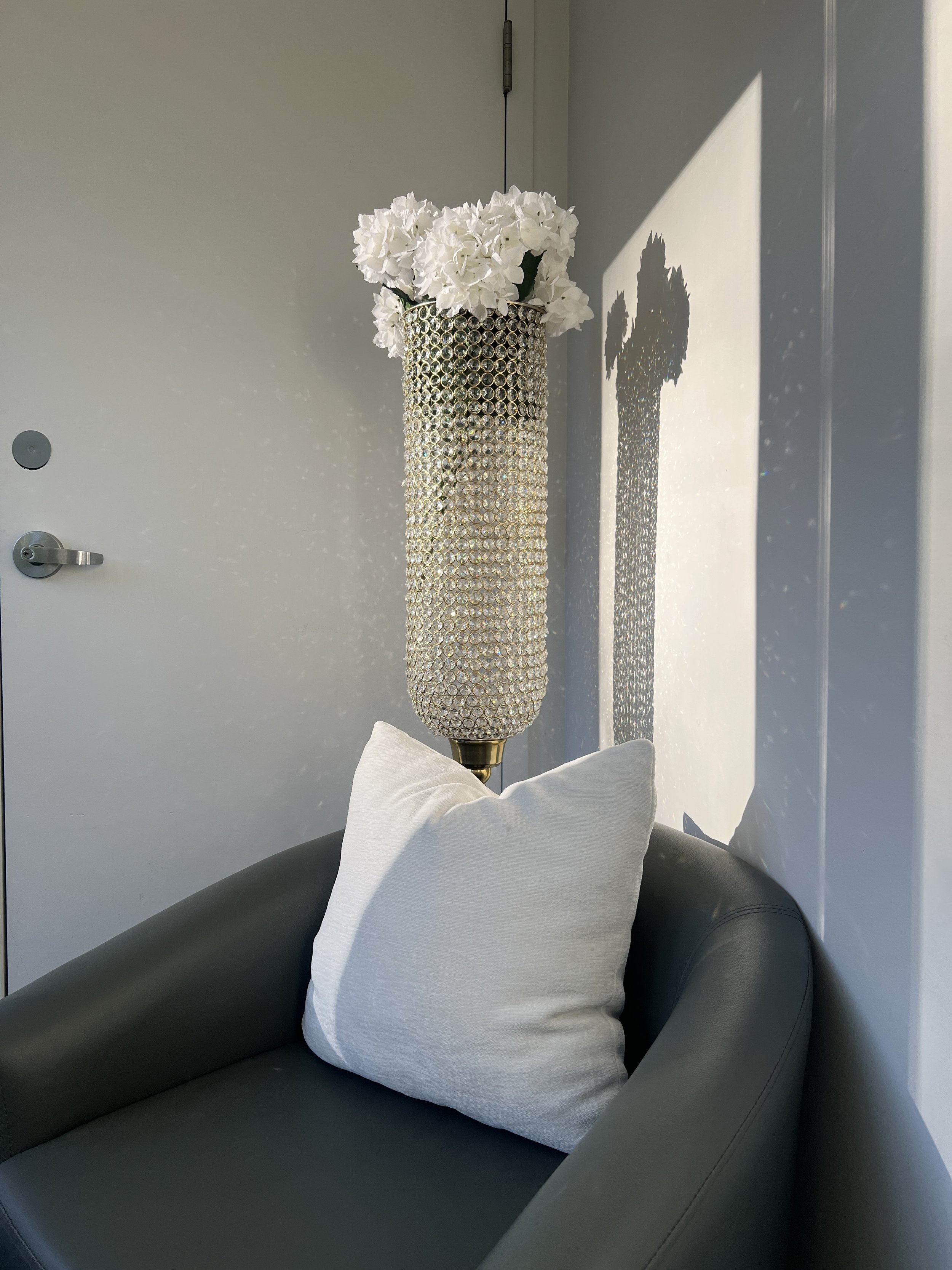 This it’s photo of a grey seat with a white pillow on it. Behind the chair  is a crystal vase holding white flowers.