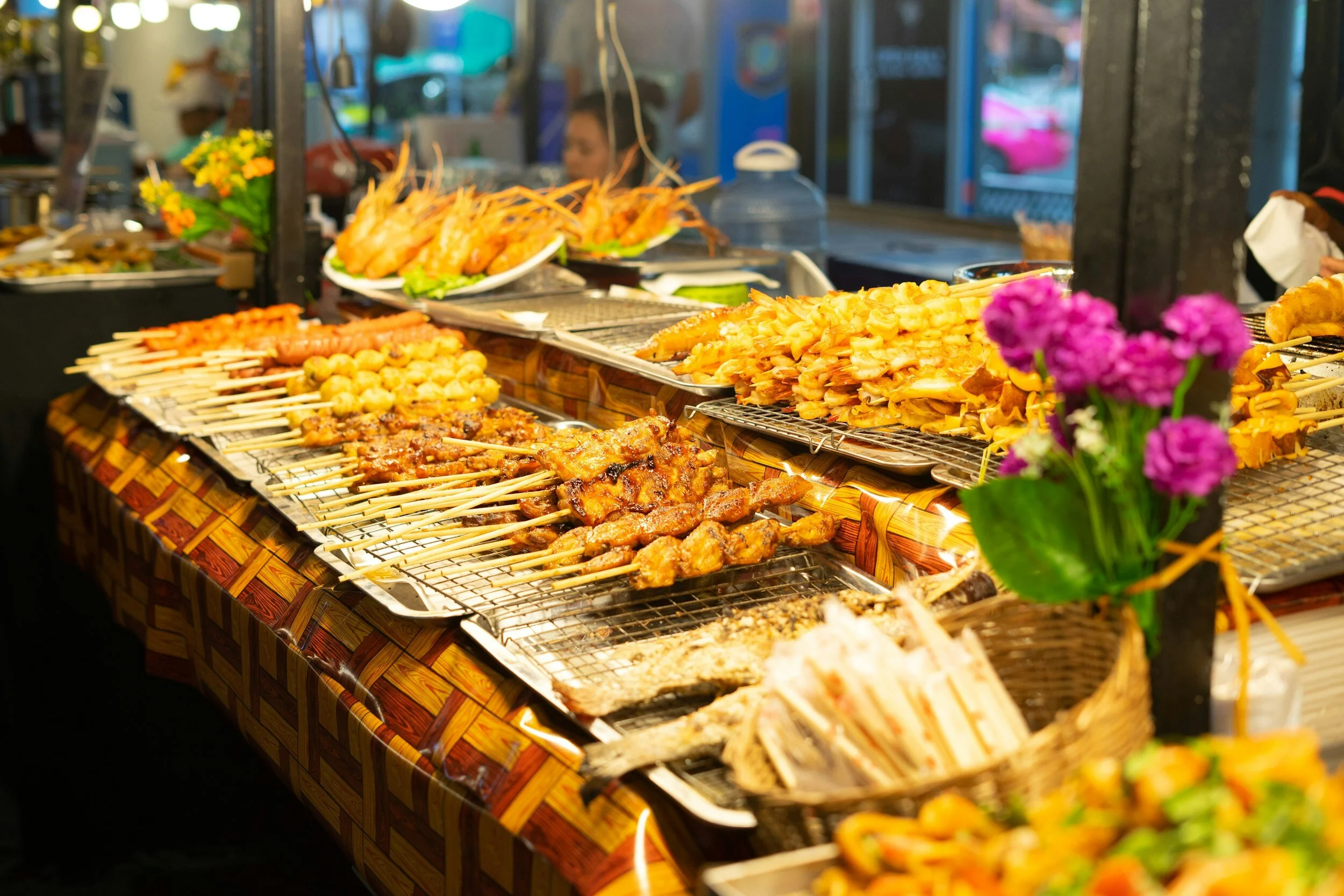 Street food stall with skewers of grilled meat and seafood on display, decorative flowers in foreground.