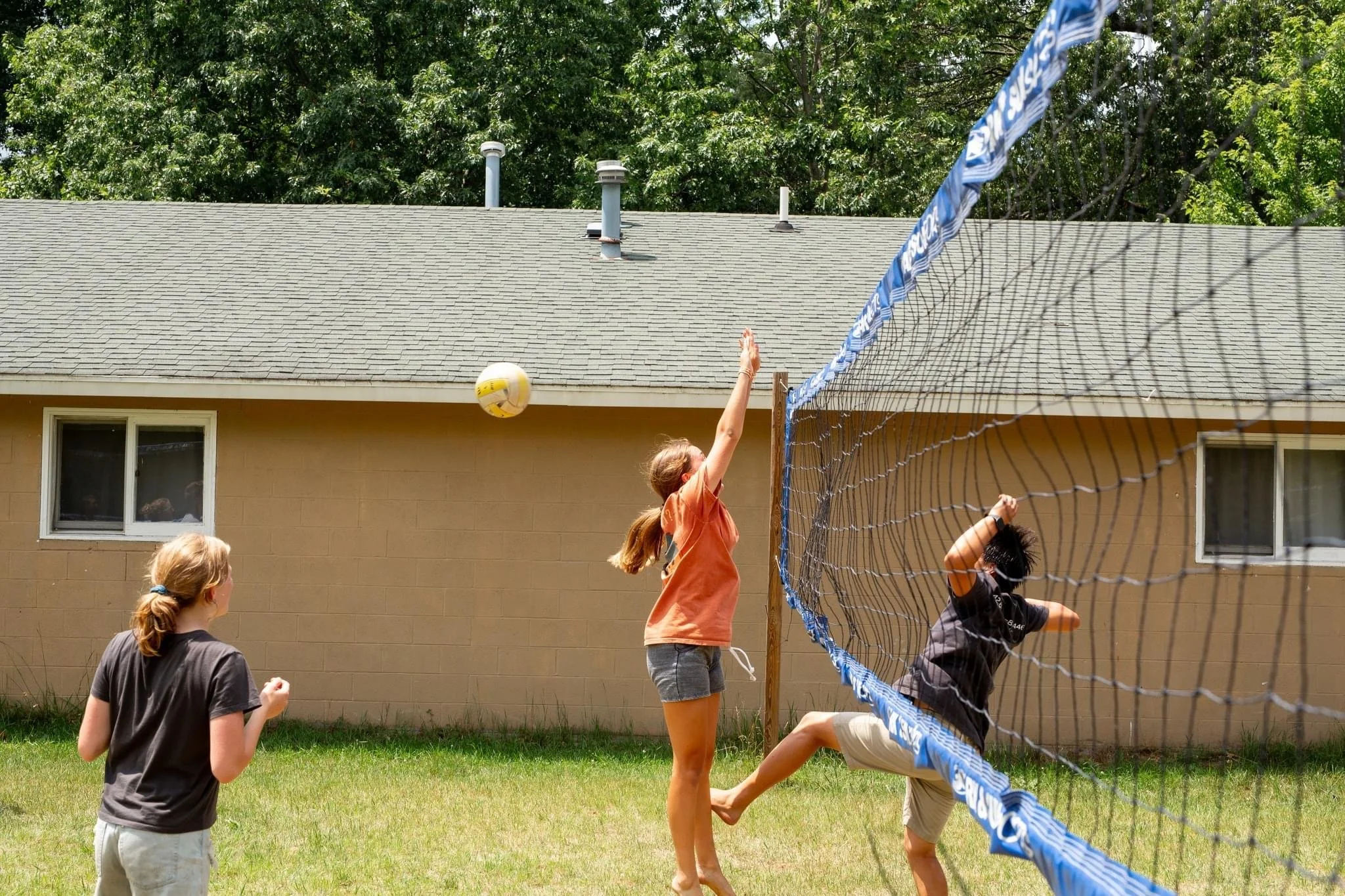 People playing volleyball in a backyard with a yellow ball, near a tan building and grass lawn.