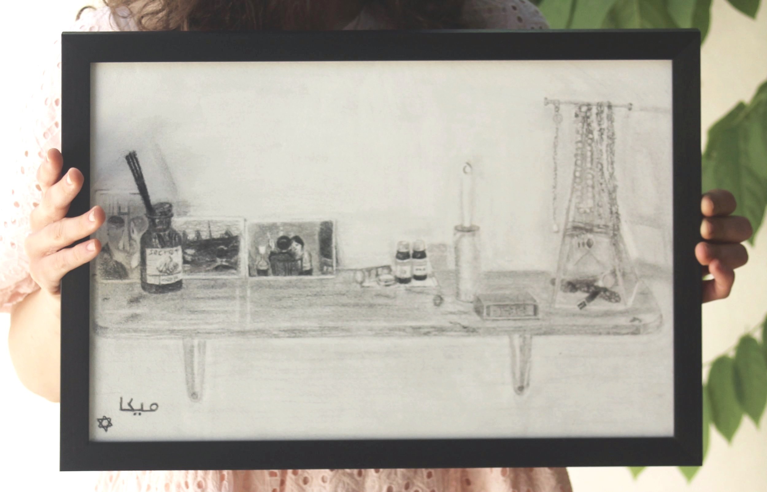 A person holding a black framed drawing of a shelf with various bottles, a candle, and objects, with writing in Arabic in the bottom left corner.