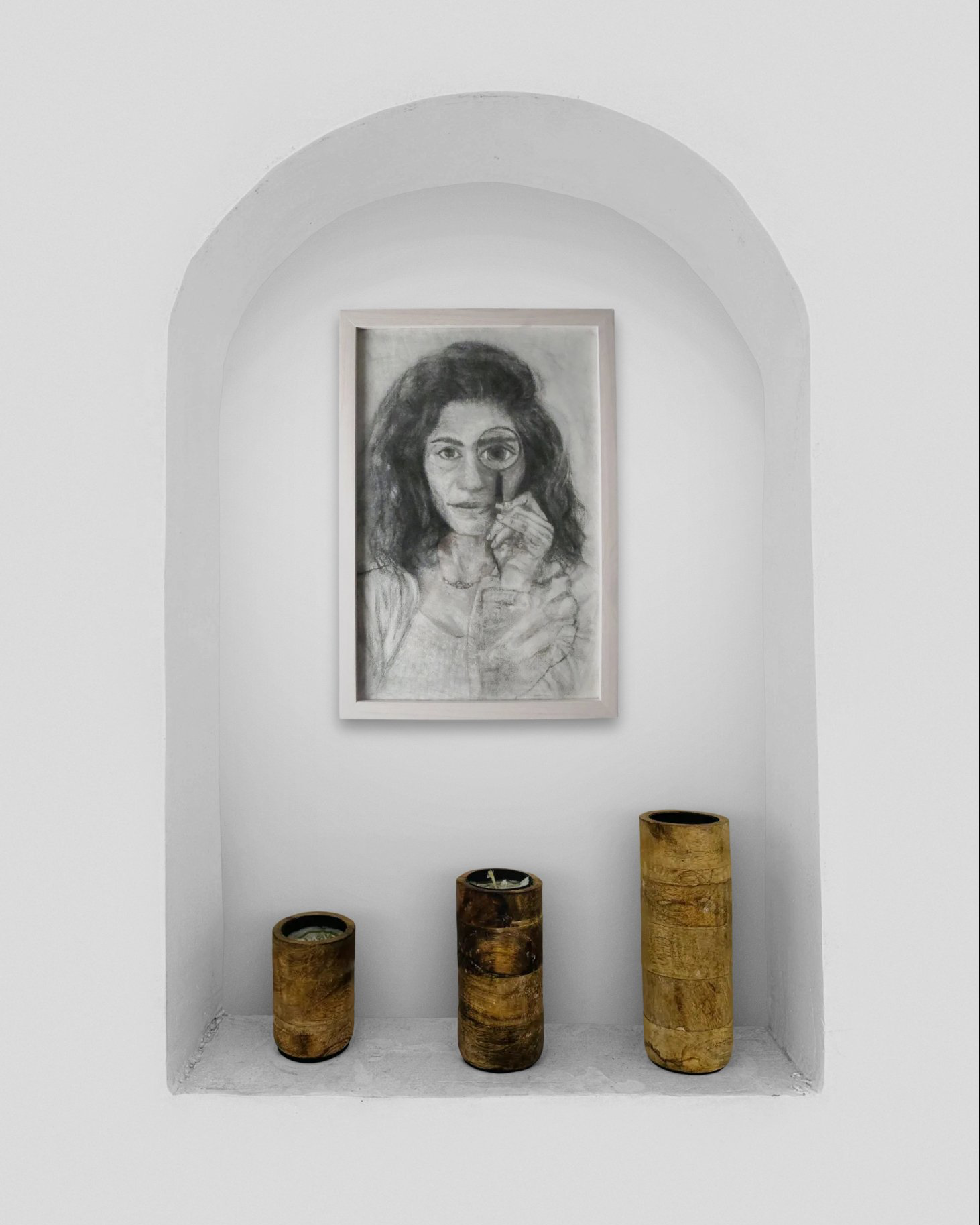 A black and white portrait of a woman with wavy hair, holding a magnifying glass, is displayed on a white wall within a recessed niche. Three wooden candle holders of varying heights are placed on the floor below the portrait.