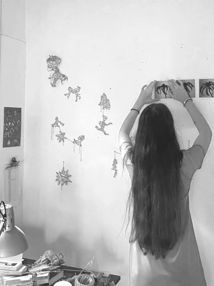 A woman with long dark hair is hanging up or arranged multiple pieces of artwork or photographs on a white wall in a room. There are various artistic images and decorations on the wall and a desk with books, a lamp, and miscellaneous items nearby.