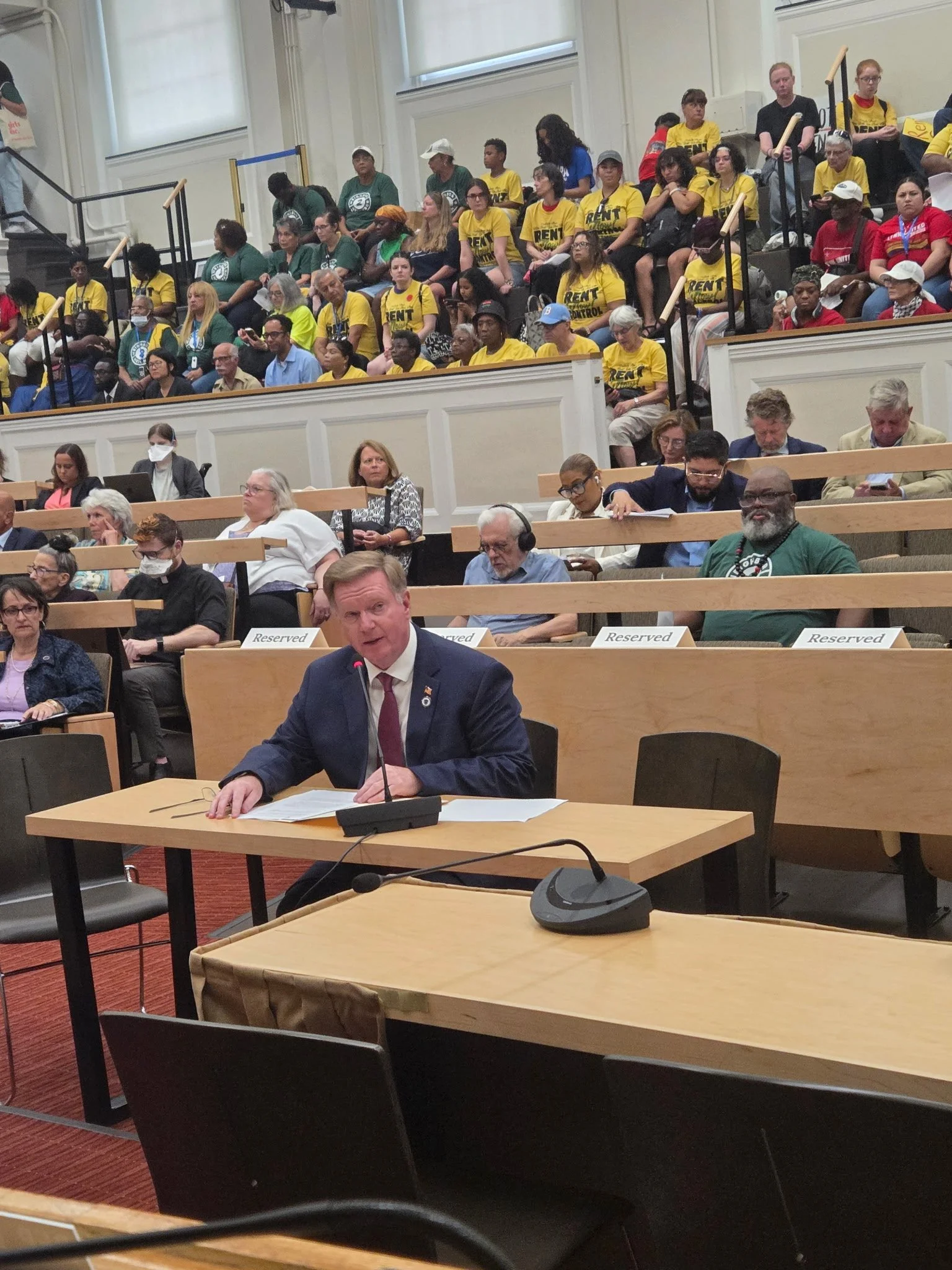 A man in a suit speaking at a table during a public hearing or meeting, with a large audience seated behind him, some wearing yellow shirts with the word 'RENT' on them.