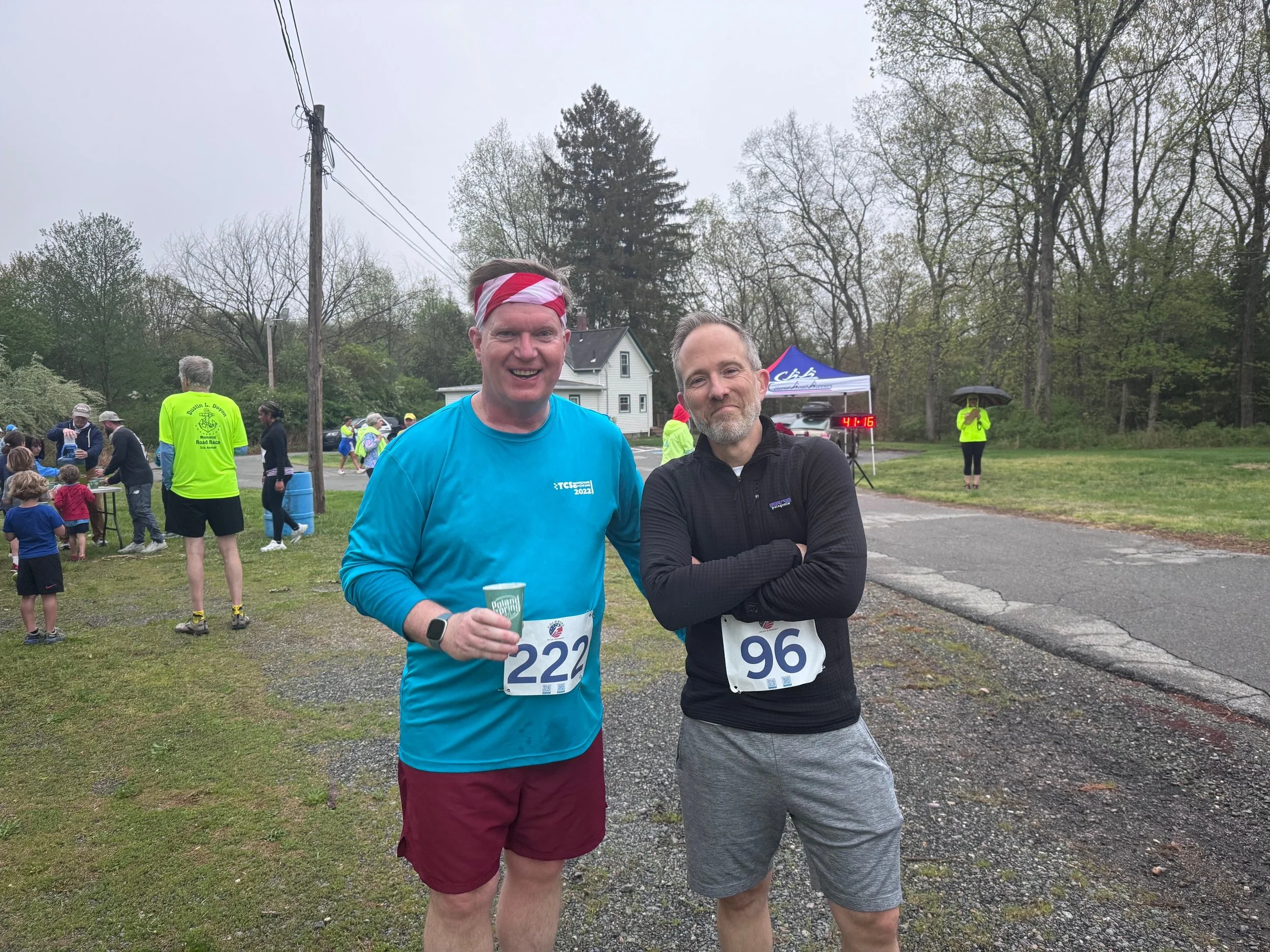 Two male runners at a race finish area, one smiling and wearing a blue shirt with bib number 222, and the other with arms crossed wearing a black jacket with bib number 96, with a group of people and race activities in the background on a cloudy day.