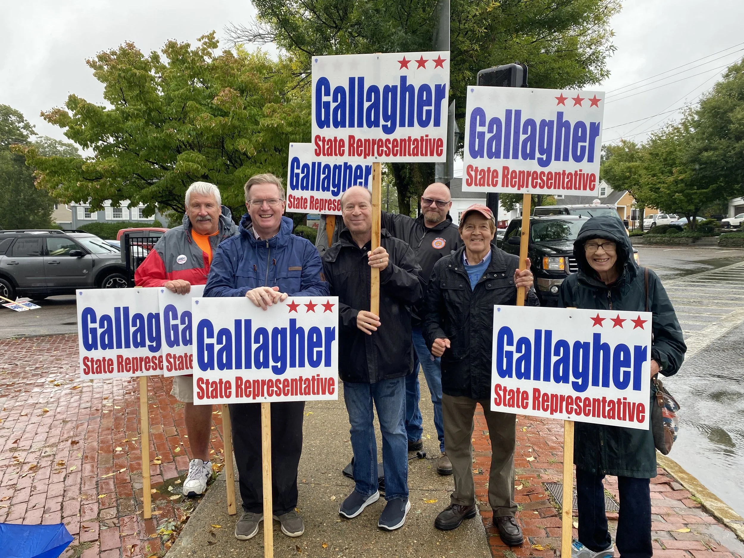 Group of people holding campaign signs for Gallagher, a state representative, on a rainy day.
