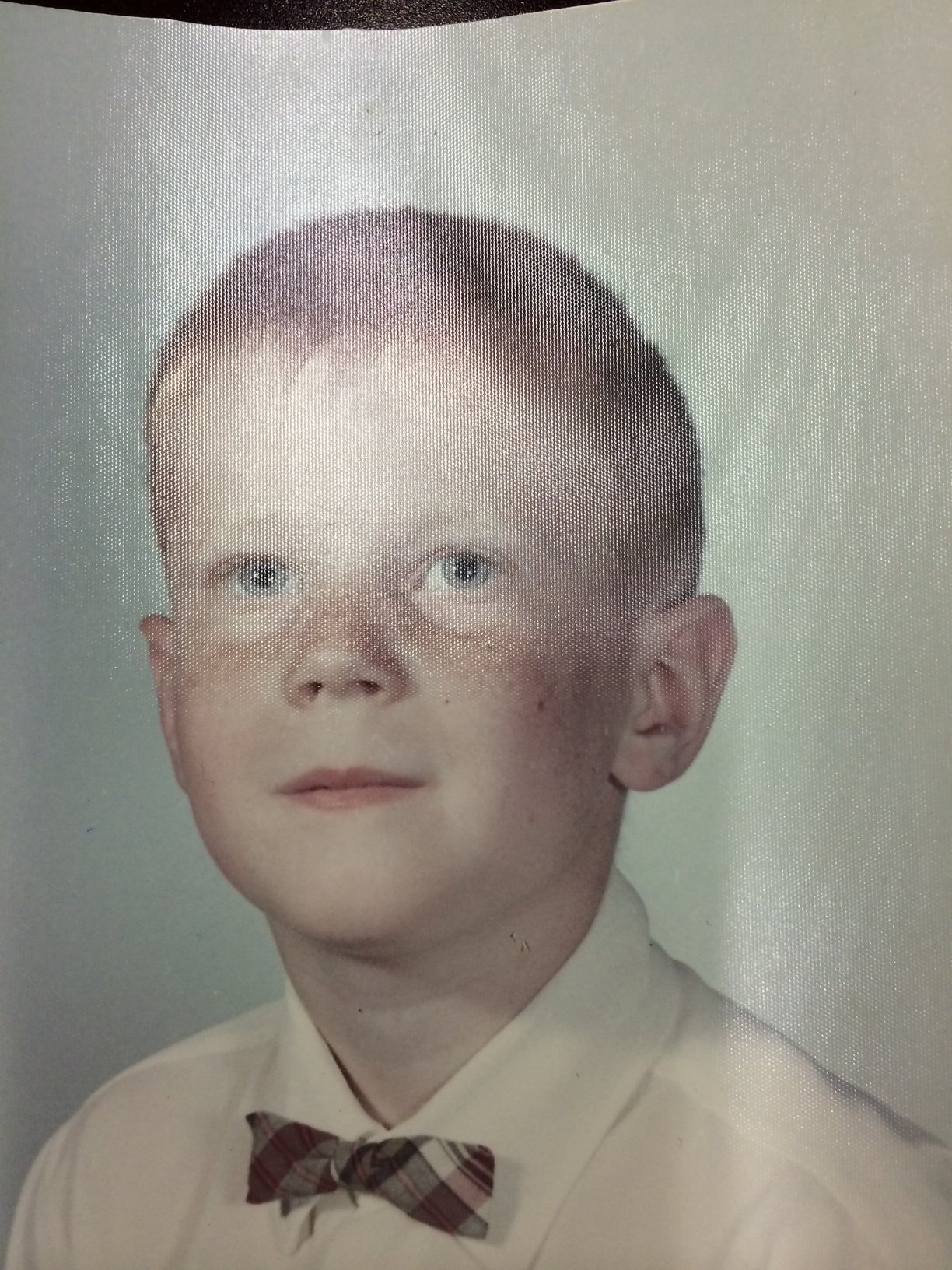 A young boy with short brown hair, blue eyes, and freckles, wearing a white shirt and a plaid bow tie, posing for a portrait against a light background.
