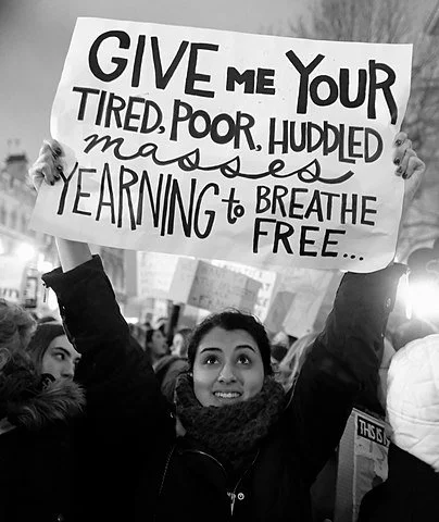 Person holding a protest sign that reads, 'Give me your tired, poor, huddled masses yearning to breathe free.'