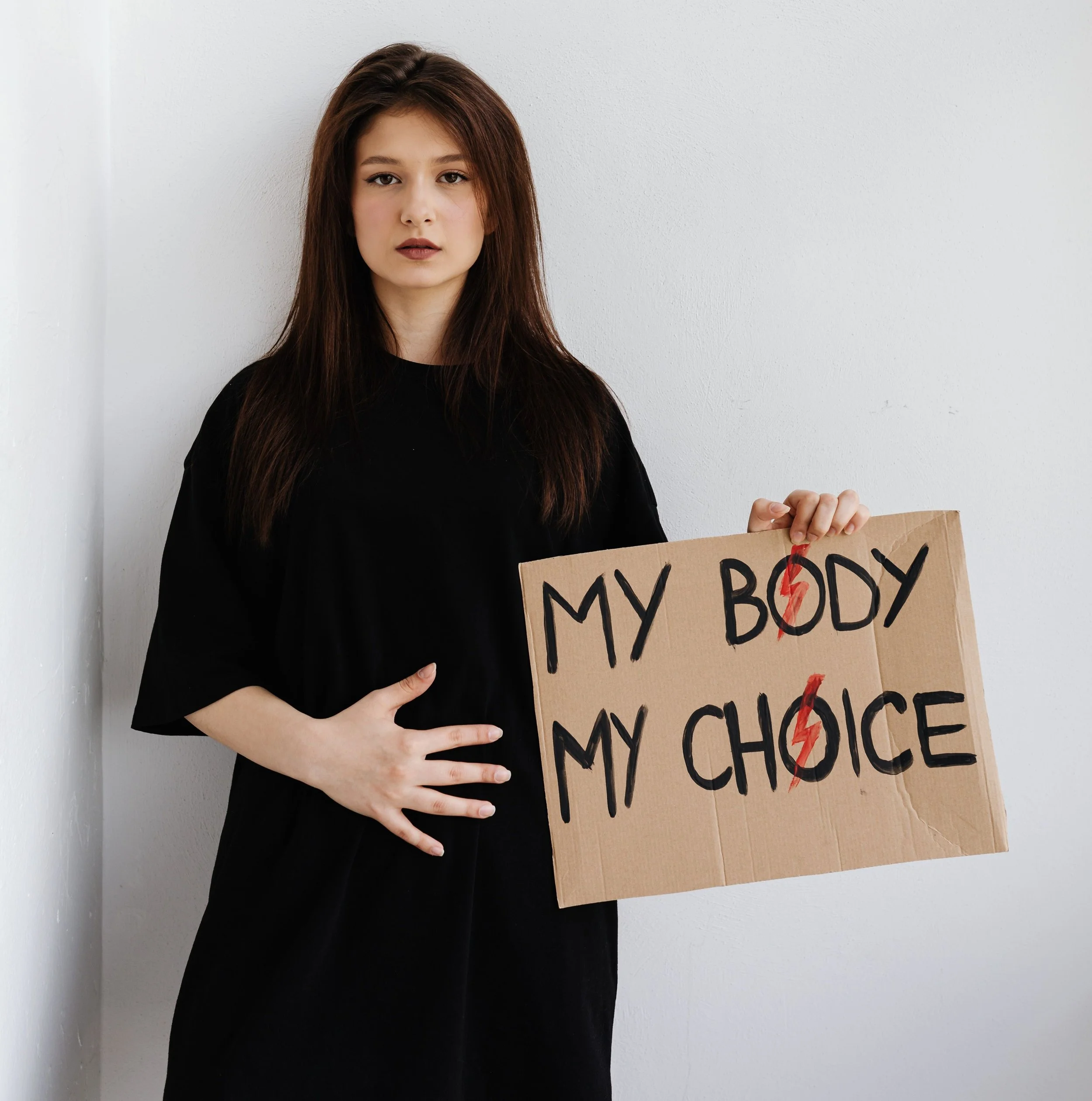 Women's Equality. Women's Rights. 14th amendment. 19th amendment. Woman with long brown hair wearing a black top holding a cardboard sign that says "MY BODY MY CHOICE" .